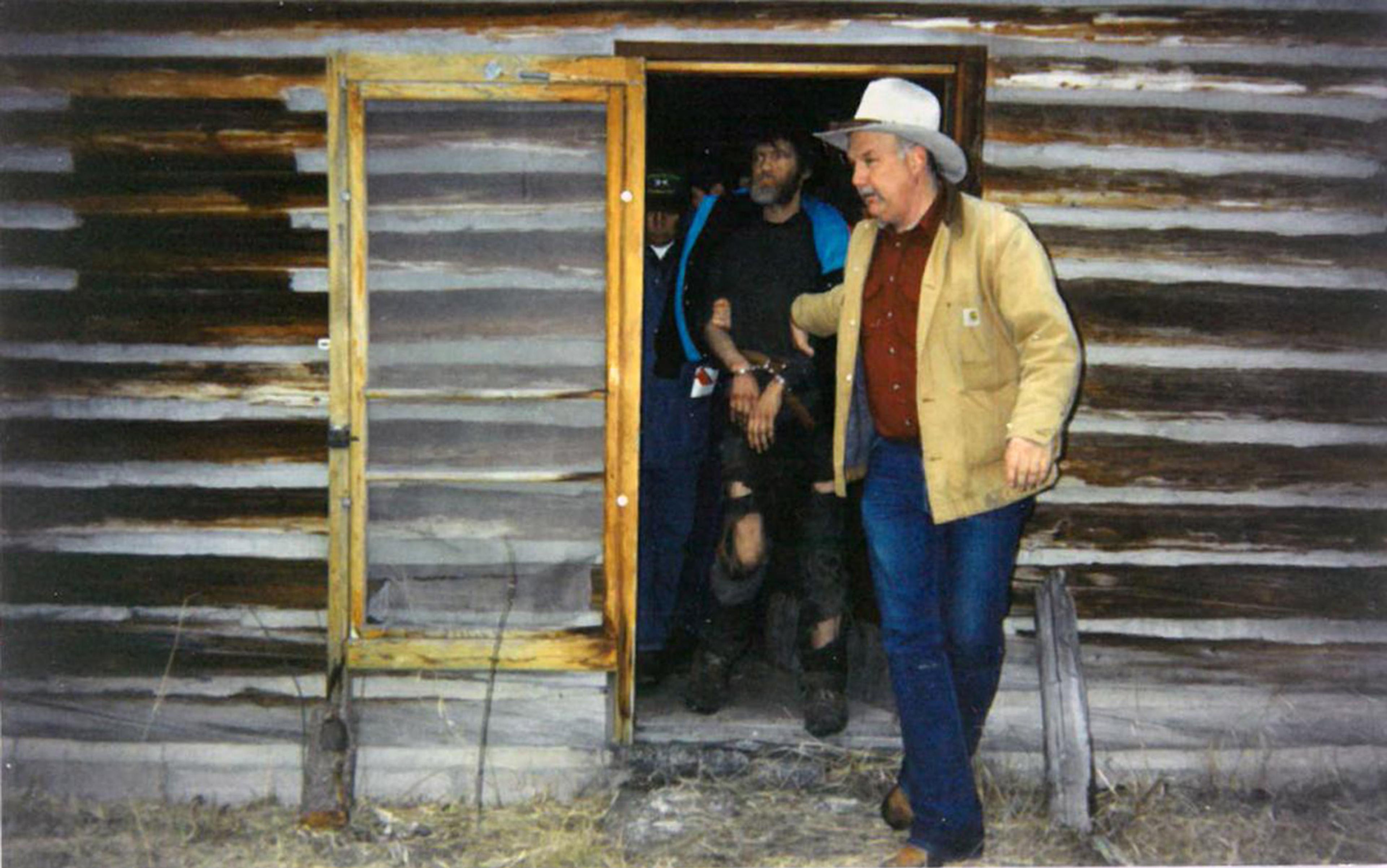 Two men escort a handcuffed man from a cabin with a wooden exterior. The men are wearing casual clothing, including a cowboy hat.