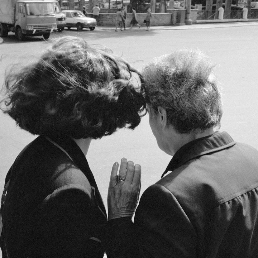 A woman seen from behind is advising another woman on something. They are standing at the edge of a wide road