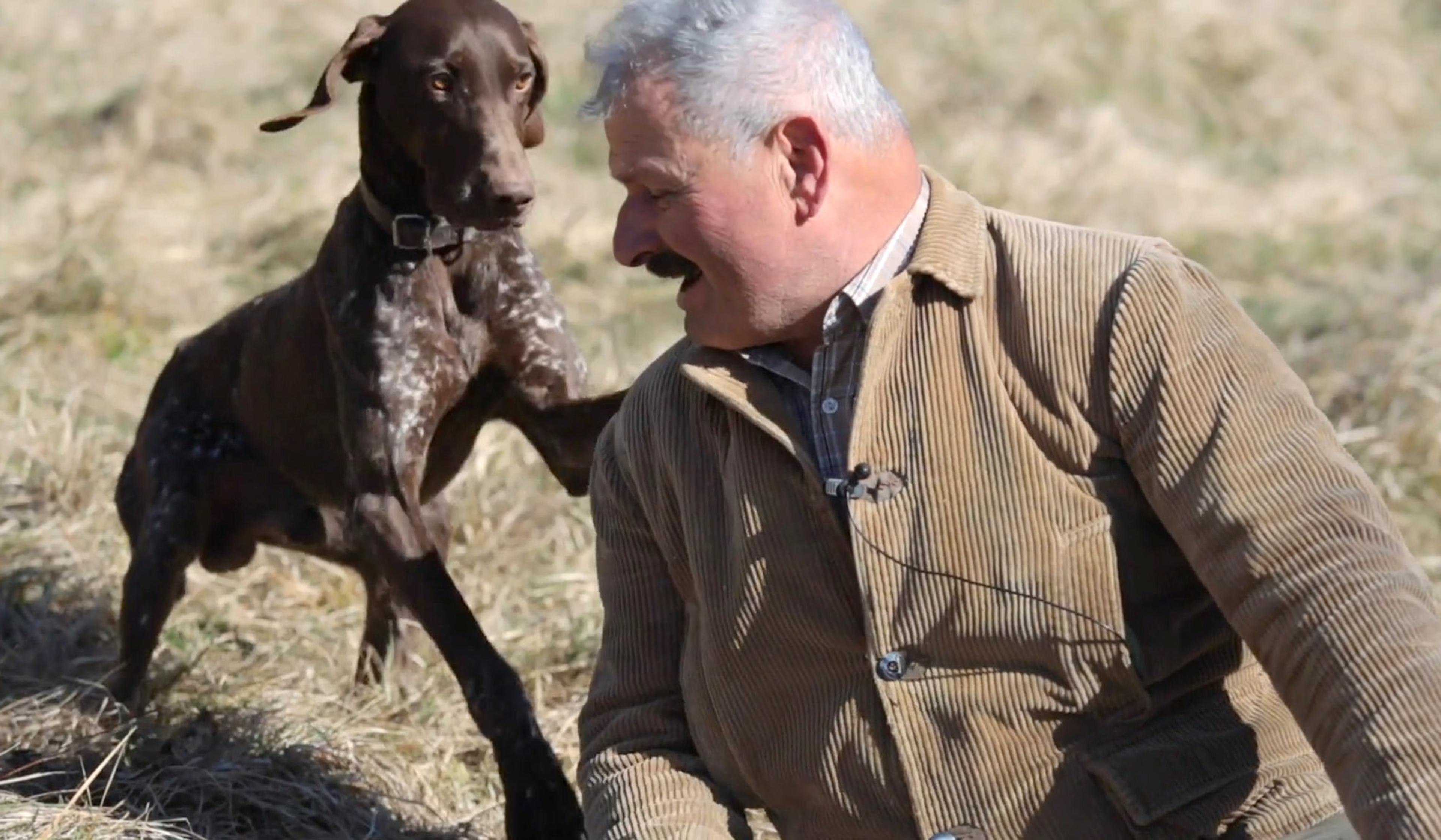 A man wearing a brown jacket and a dog in a field. The dog appears to be moving towards the man who is looking at it.