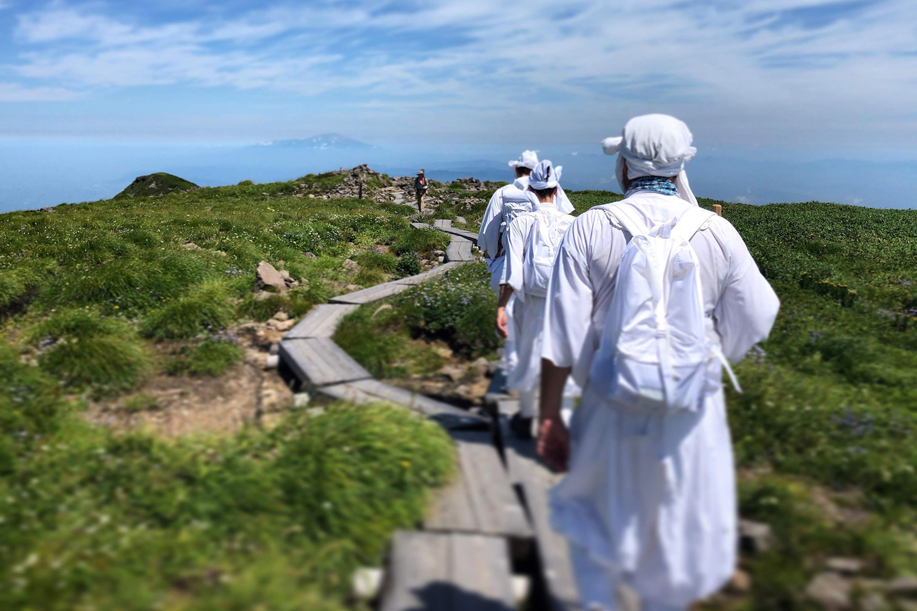 A group of people in white attire and backpacks walk along a wooden path through green fields under a blue sky with scattered clouds.