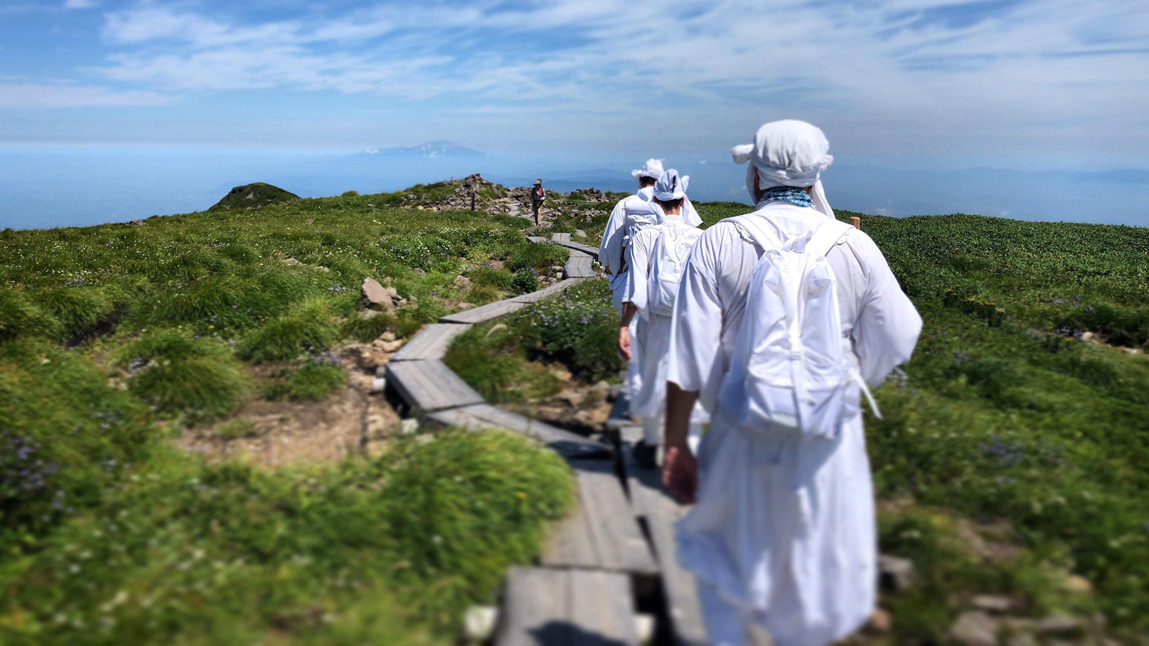 A group of people in white attire and backpacks walk along a wooden path through green fields under a blue sky with scattered clouds.