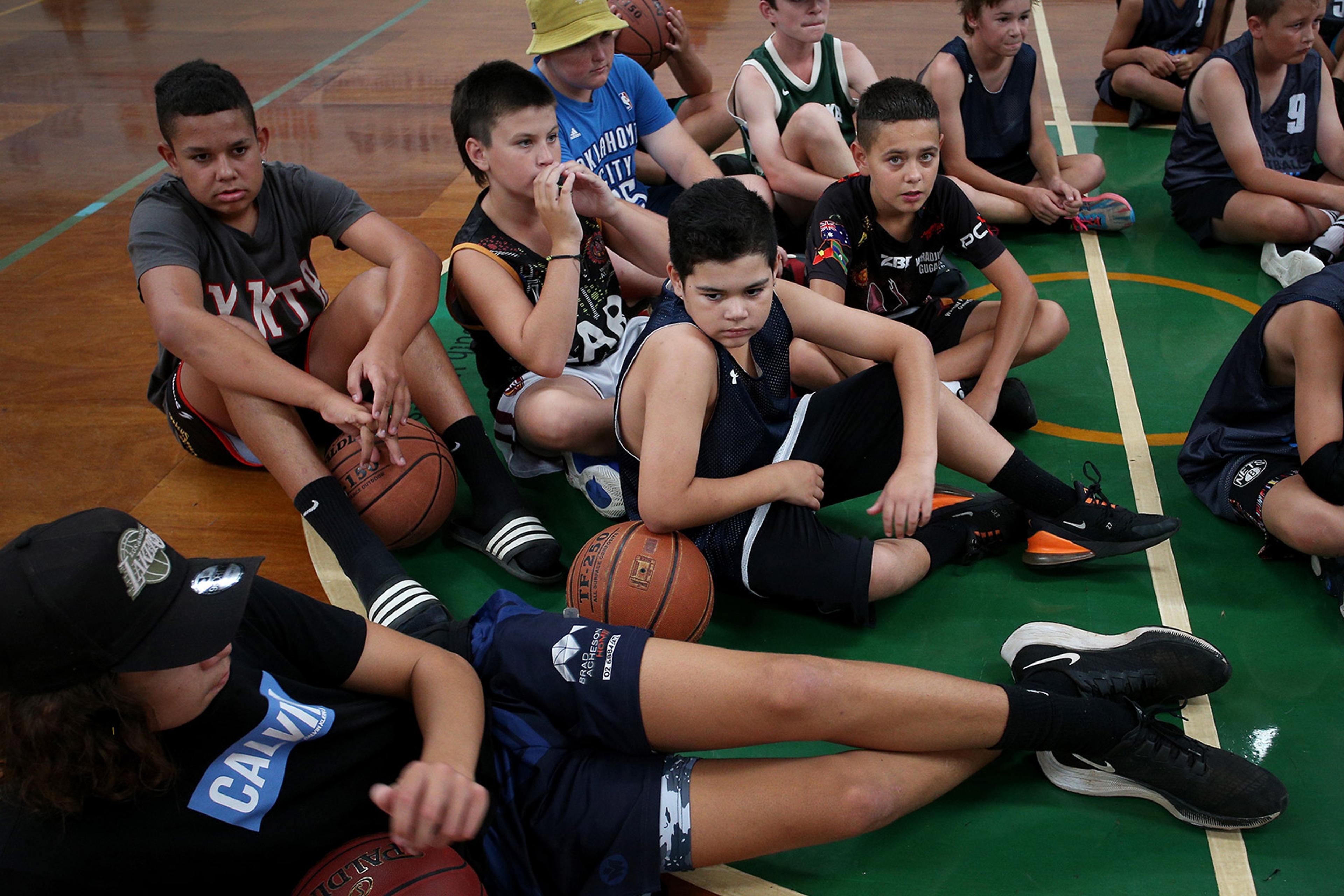 A group of children sitting on a gymnasium floor, holding basketballs, wearing various athletic clothing, and appearing to be in a sports class.