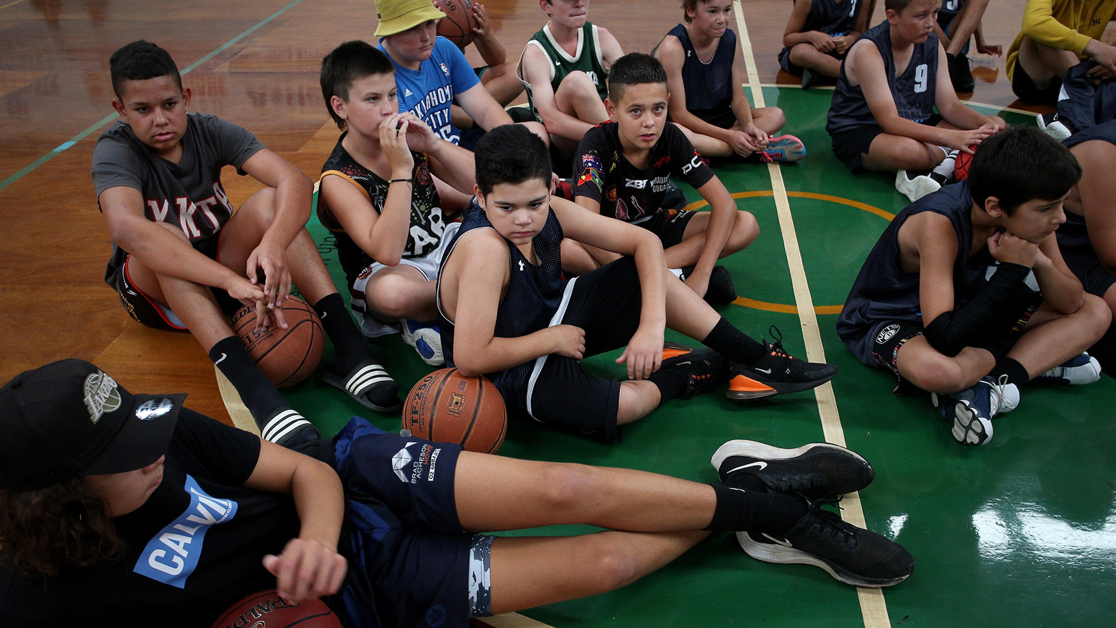 A group of children sitting on a gymnasium floor, holding basketballs, wearing various athletic clothing, and appearing to be in a sports class.