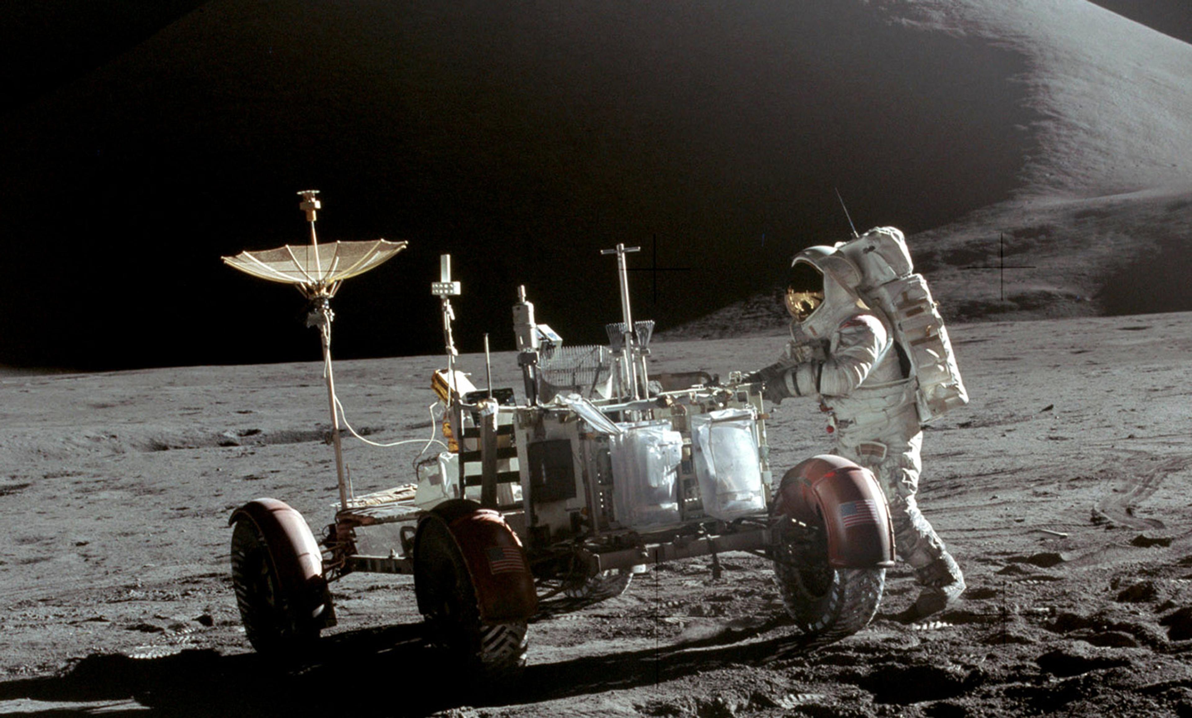 An astronaut in a space suit standing next to a lunar rover on the Moon’s surface with mountains in the background.