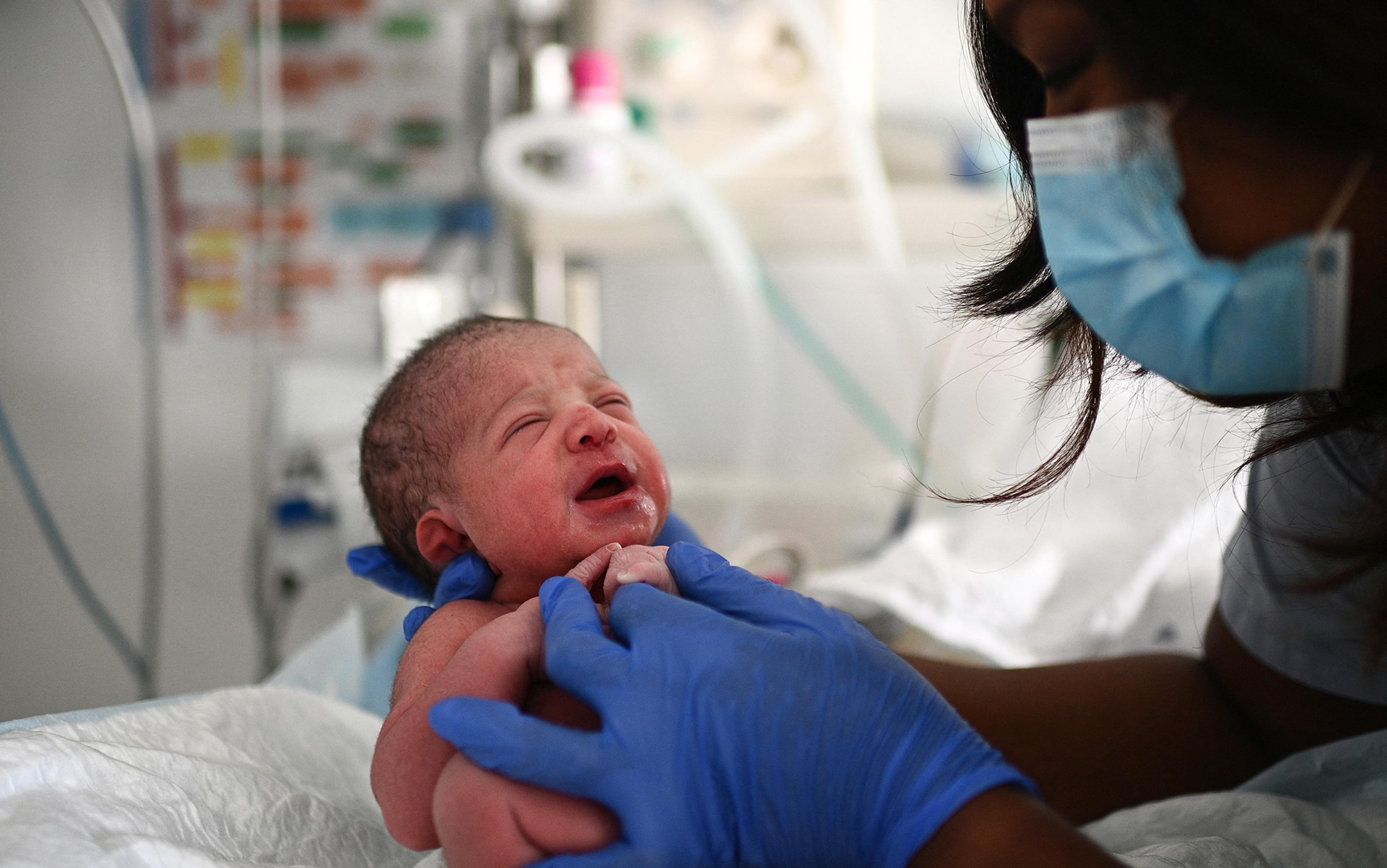 Newborn baby being held by a person wearing blue gloves, with another masked individual looking at the baby in a medical setting.