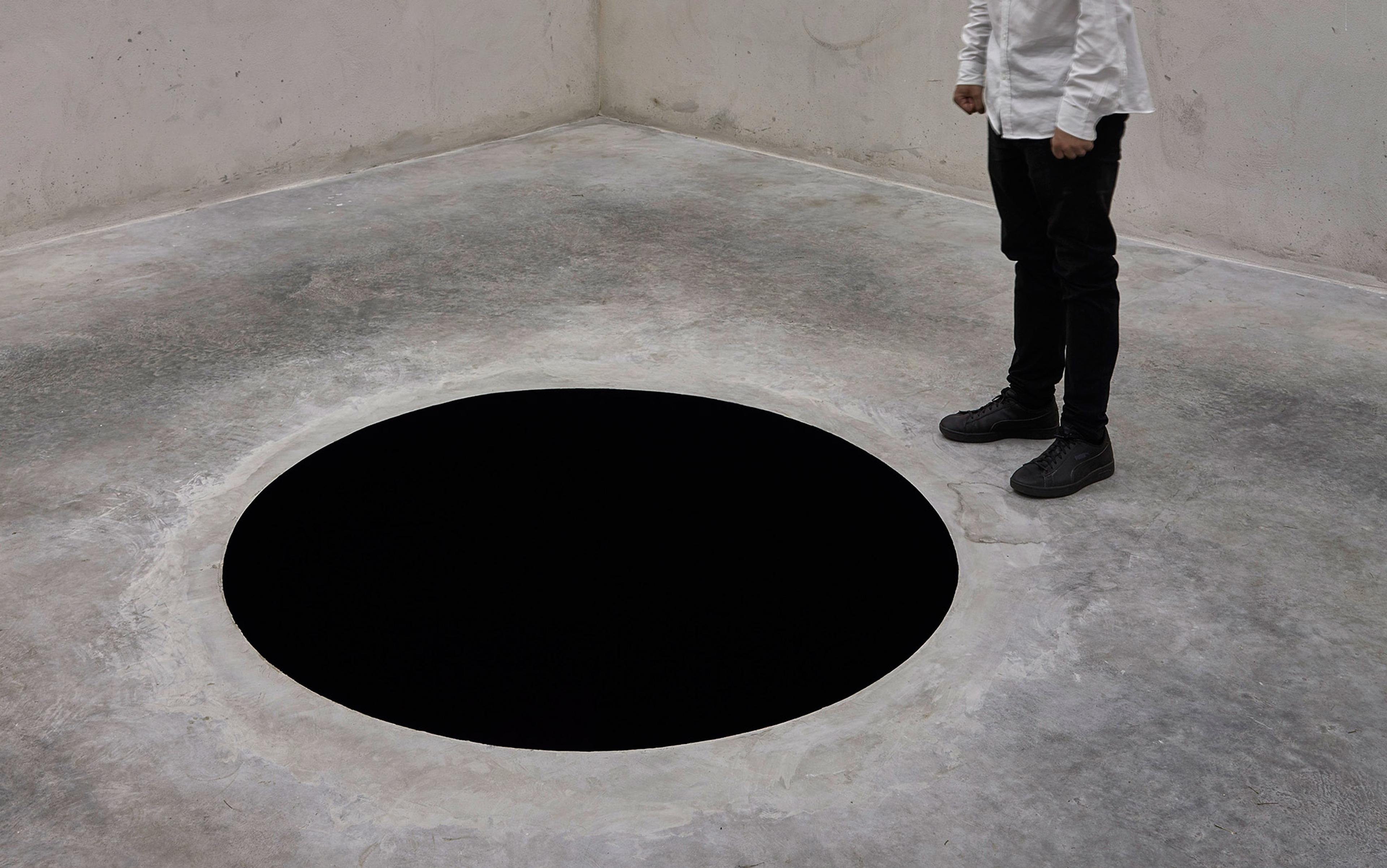 A person standing near a large black circular void in the floor of a grey, minimalist room.