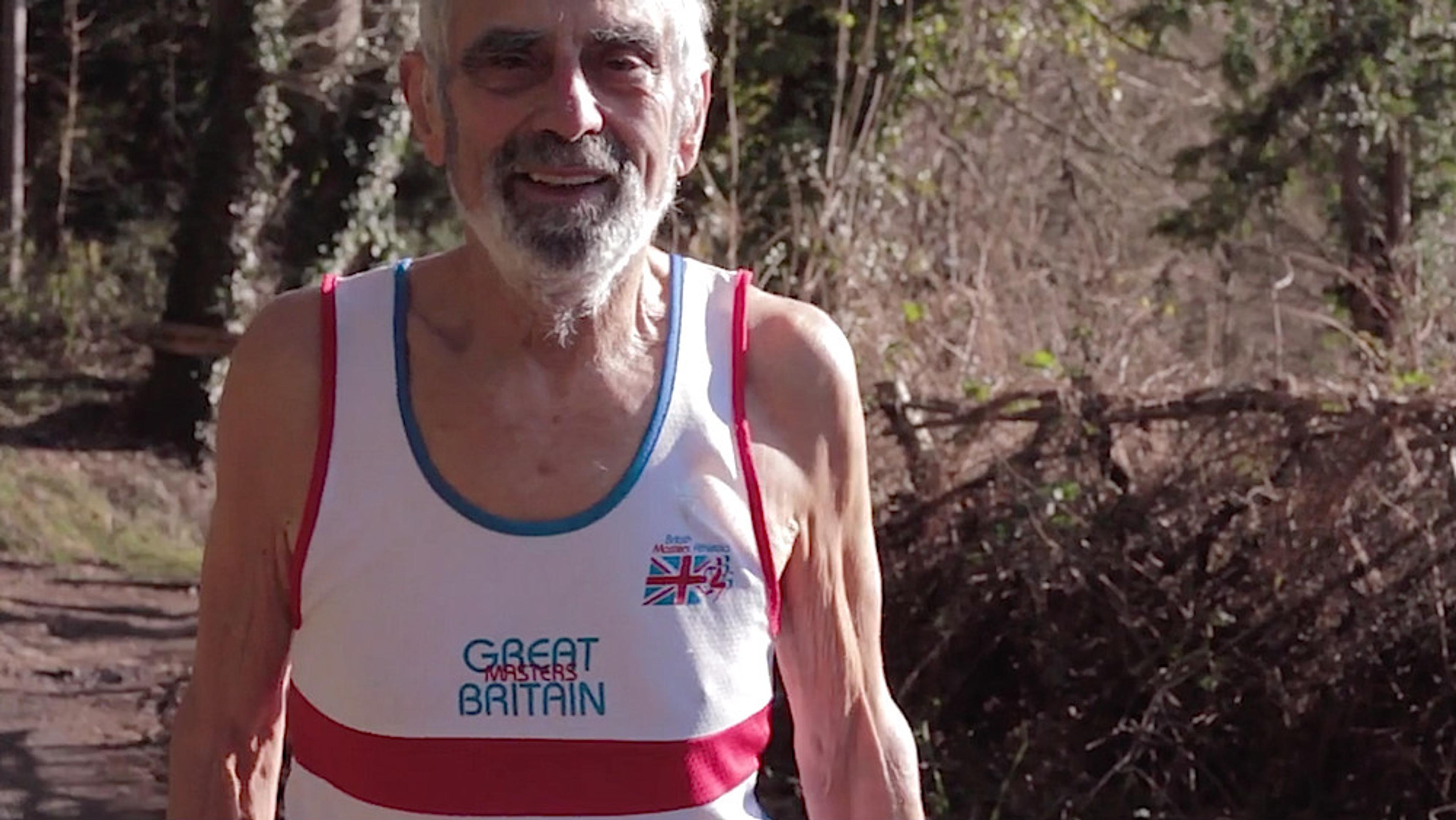 An elderly man outdoors smiling, wearing a Great Masters Britain running vest with a British flag, trees in the background.
