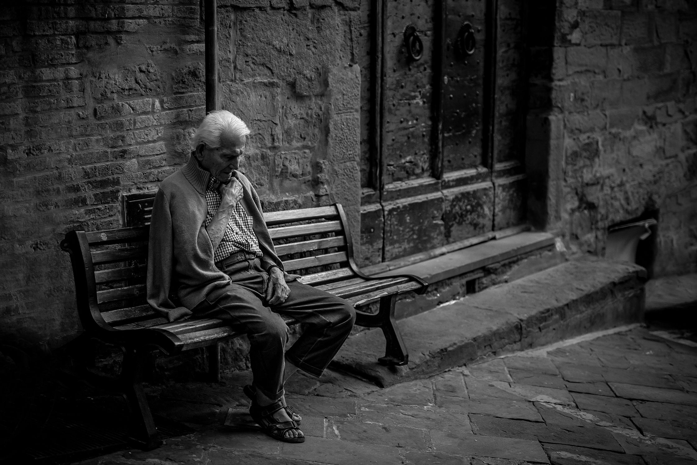 Black and white photo of an elderly man sitting on a bench in a cobblestone street, wearing a cardigan and sandals.