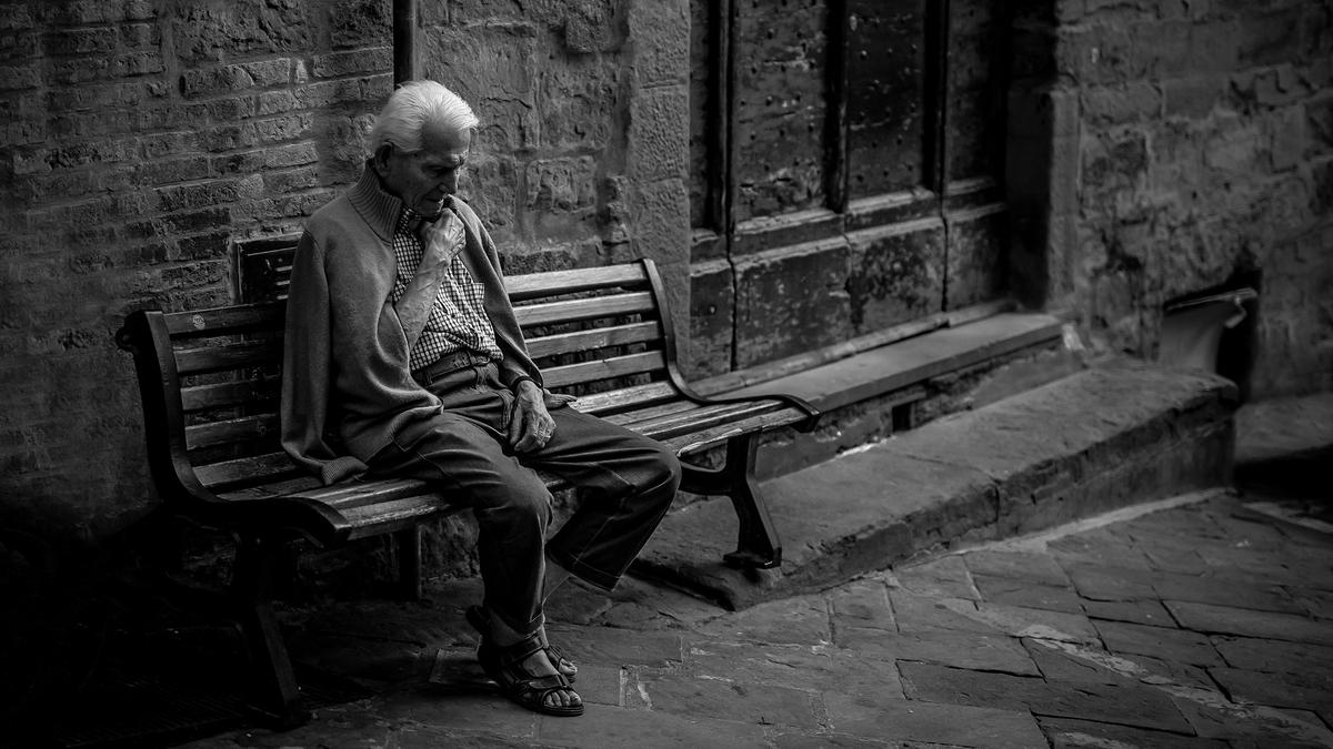 Black and white photo of an elderly man sitting on a bench in a cobblestone street, wearing a cardigan and sandals.