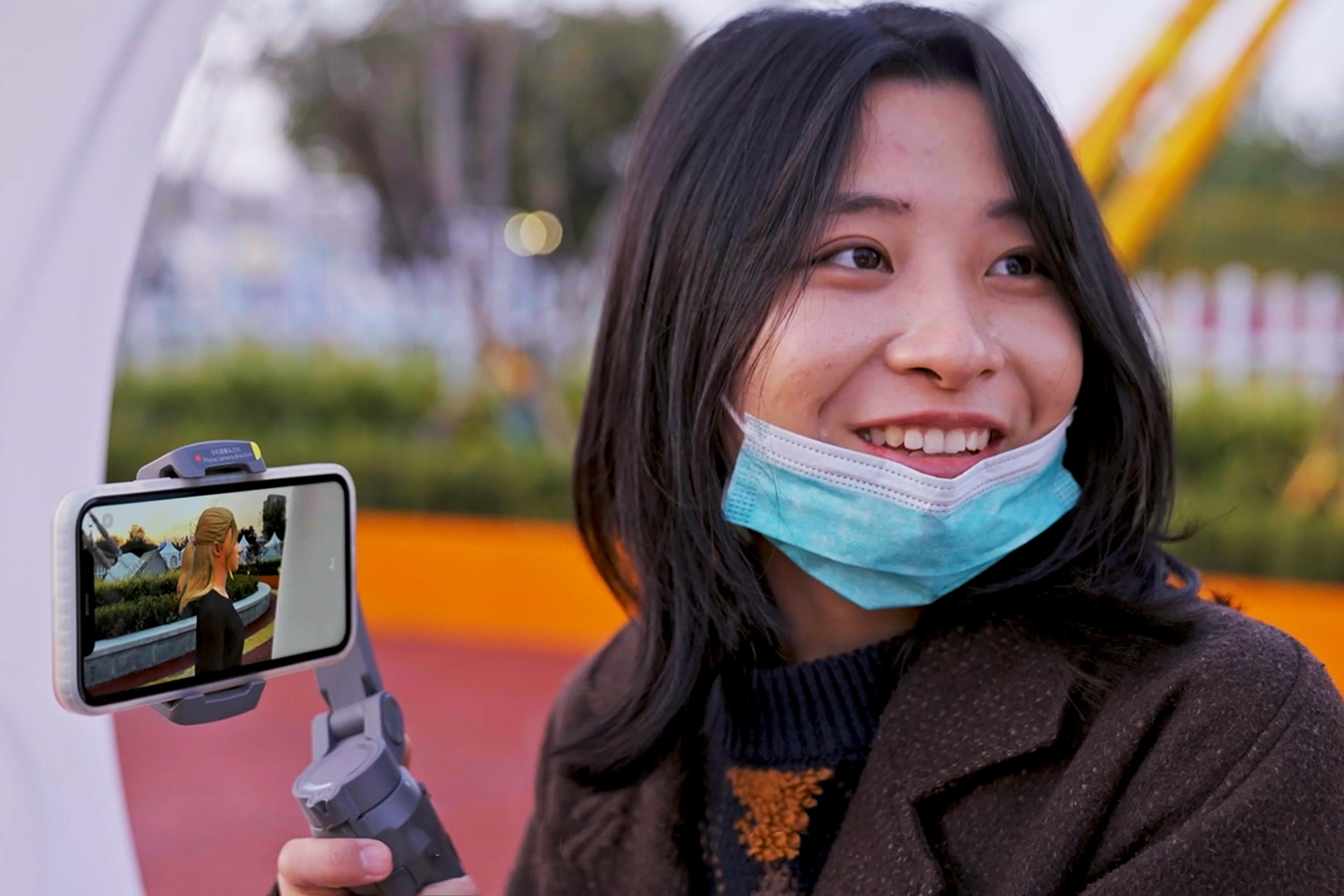 Woman outdoors smiling and holding a gimbal with mounted smartphone, showing a blonde AI woman on the phone screen, wearing a surgical mask pulled below her chin.