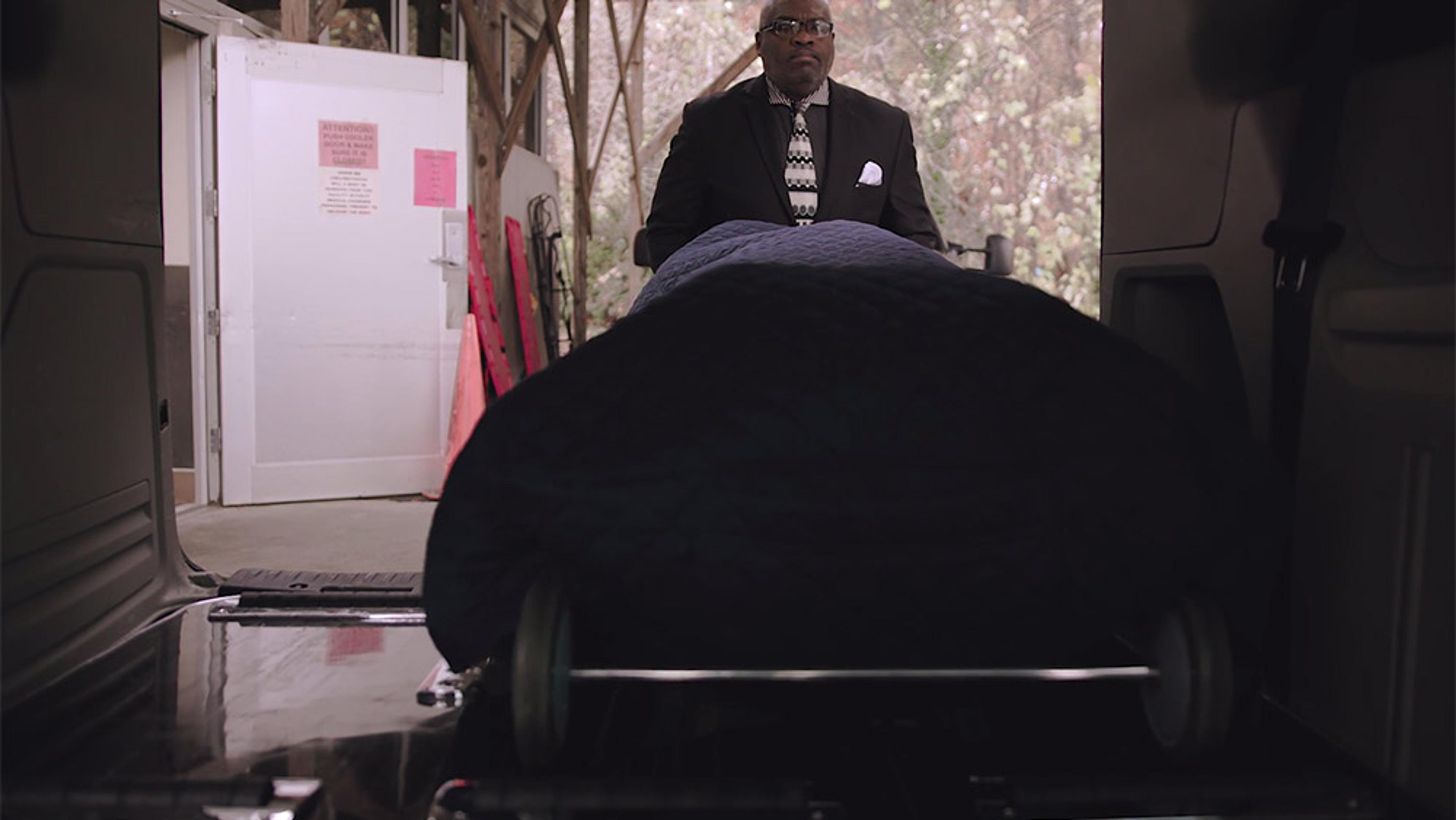 A funeral director in a suit and tie pushes a covered body on a stretcher into a vehicle. The scene is solemn.