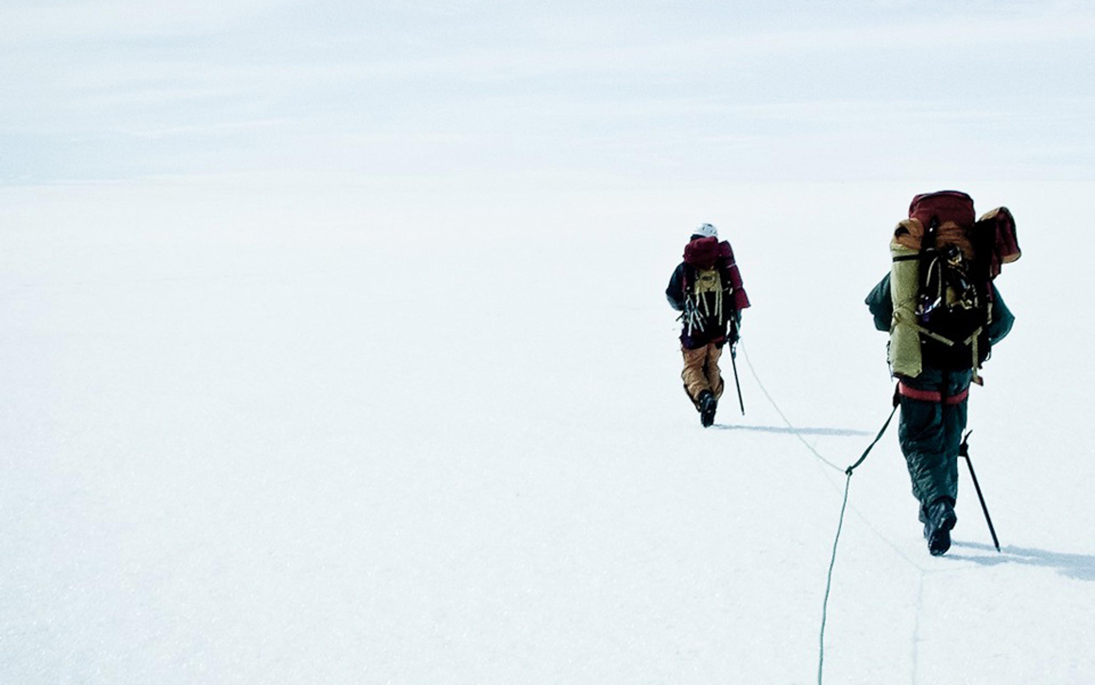 Two hikers with large backpacks walking on a vast snowy landscape, connected by a rope, heading into the distance.