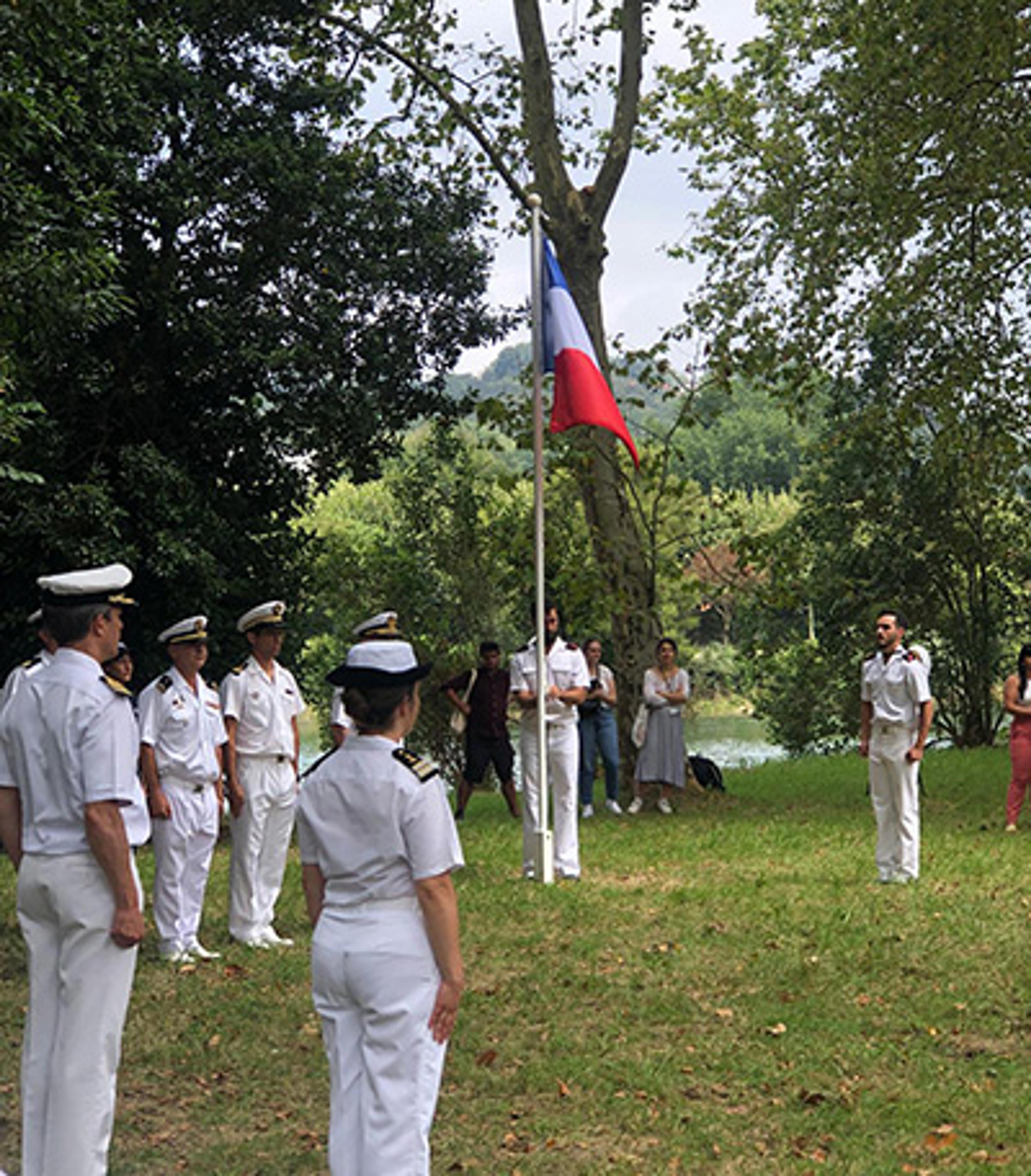 Photo of people in naval uniforms gathered around a flagpole with a French flag on a grassy area surrounded by trees.