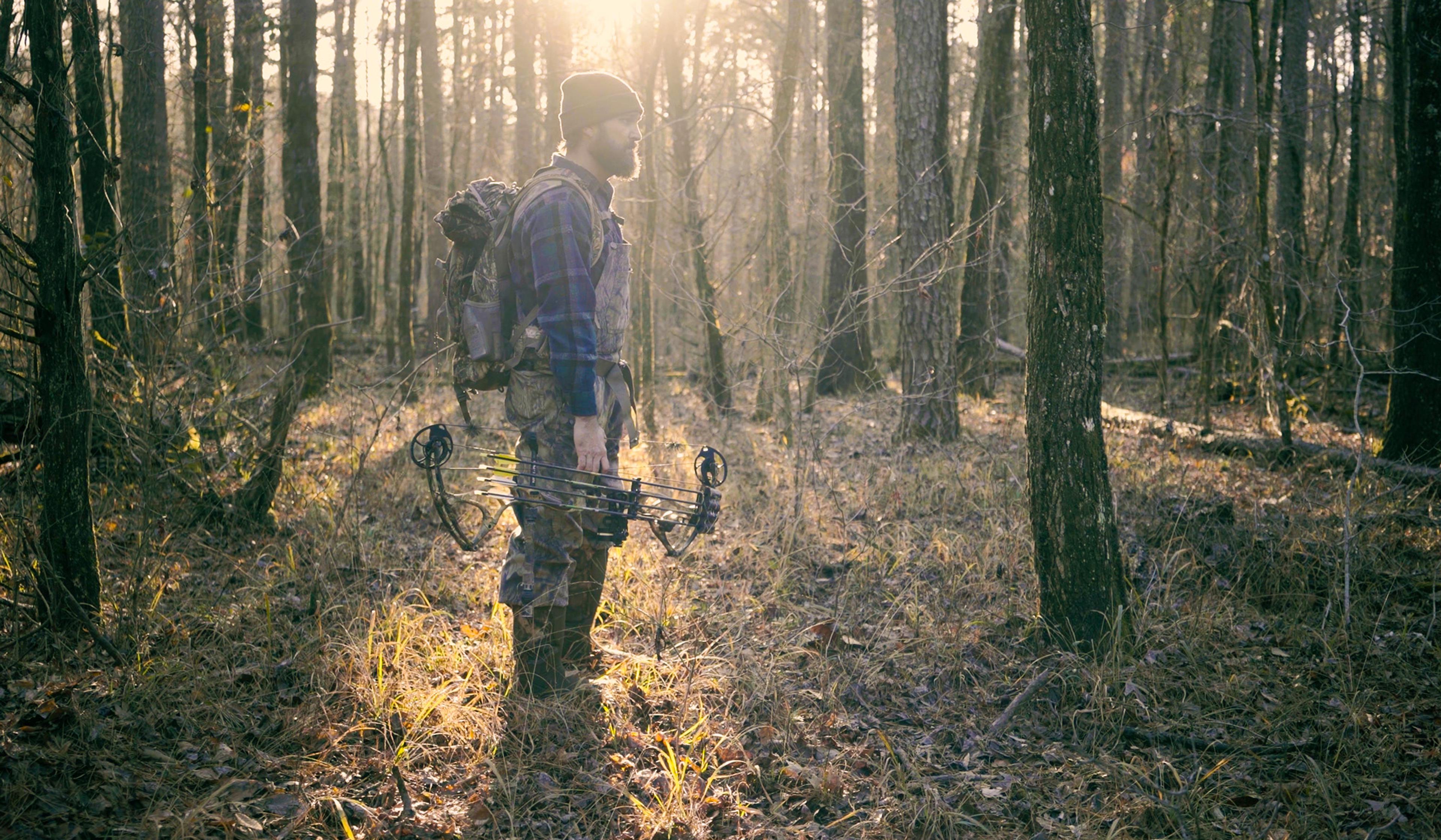 A person stands in a sunlit forest, holding a compound bow, and wearing outdoor gear with a backpack.