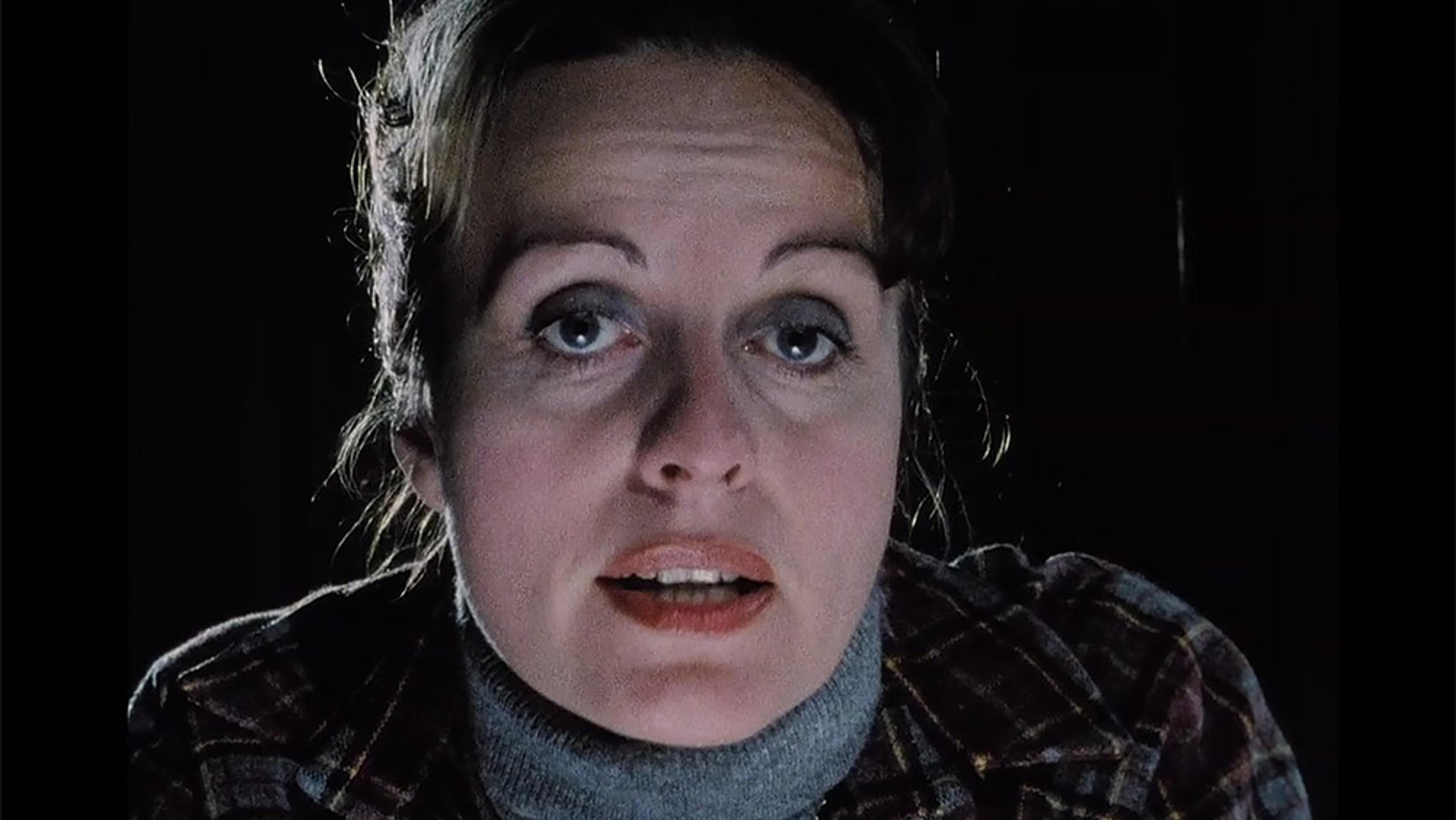 A close-up photo of a woman with light skin and blue eyes wearing a plaid shirt and a grey turtleneck, looking towards the camera.