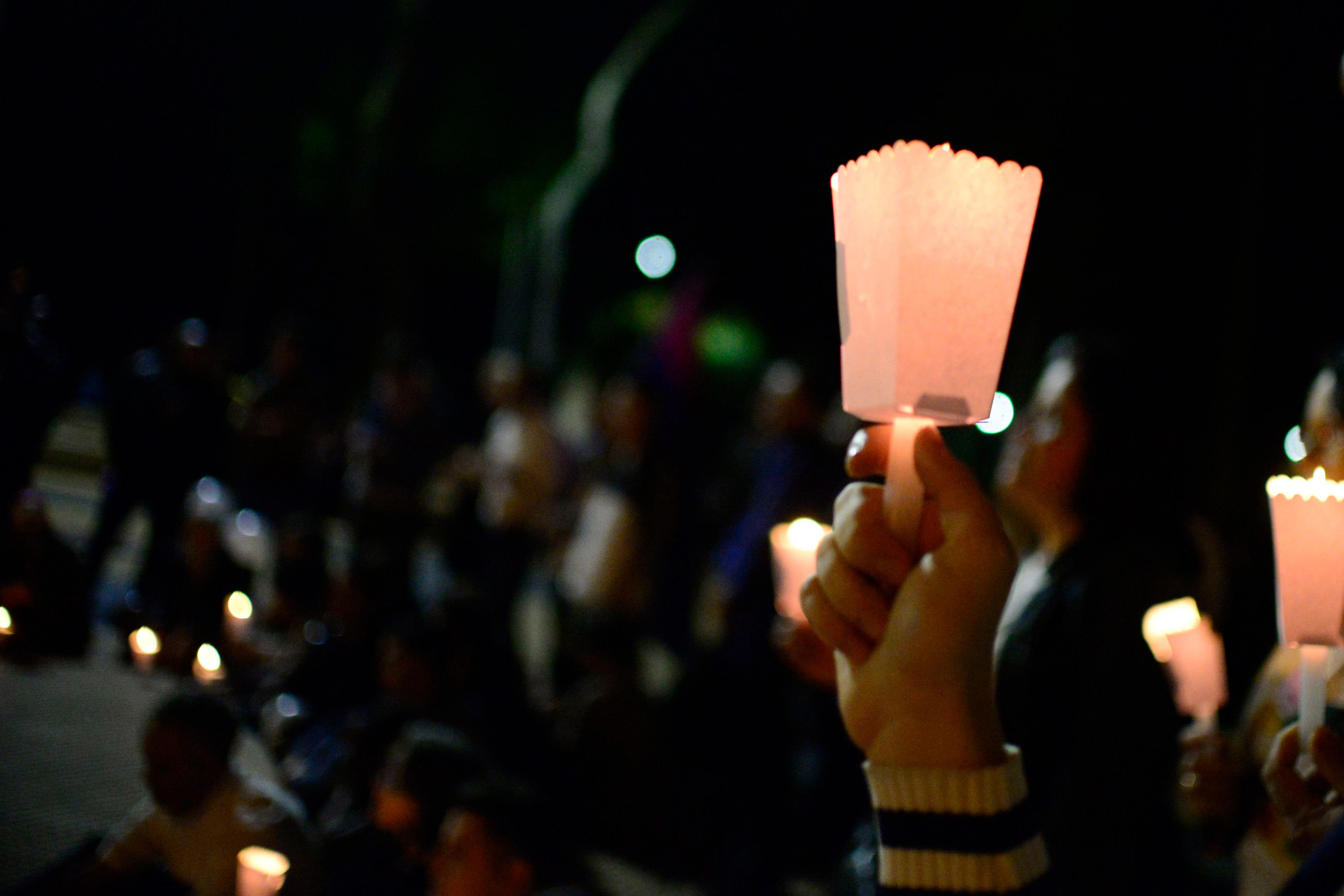Photo of a hand holding a lit candle in a paper holder at a night vigil, with blurred crowd holding candles in the background.