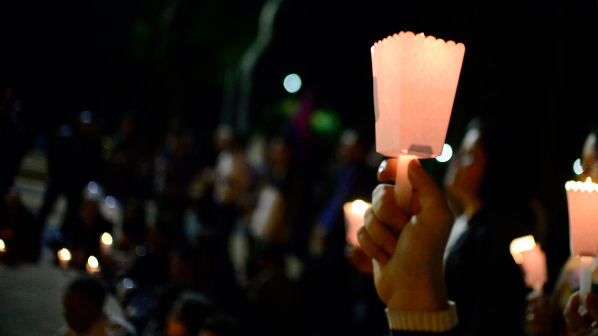 Photo of a hand holding a lit candle in a paper holder at a night vigil, with blurred crowd holding candles in the background.