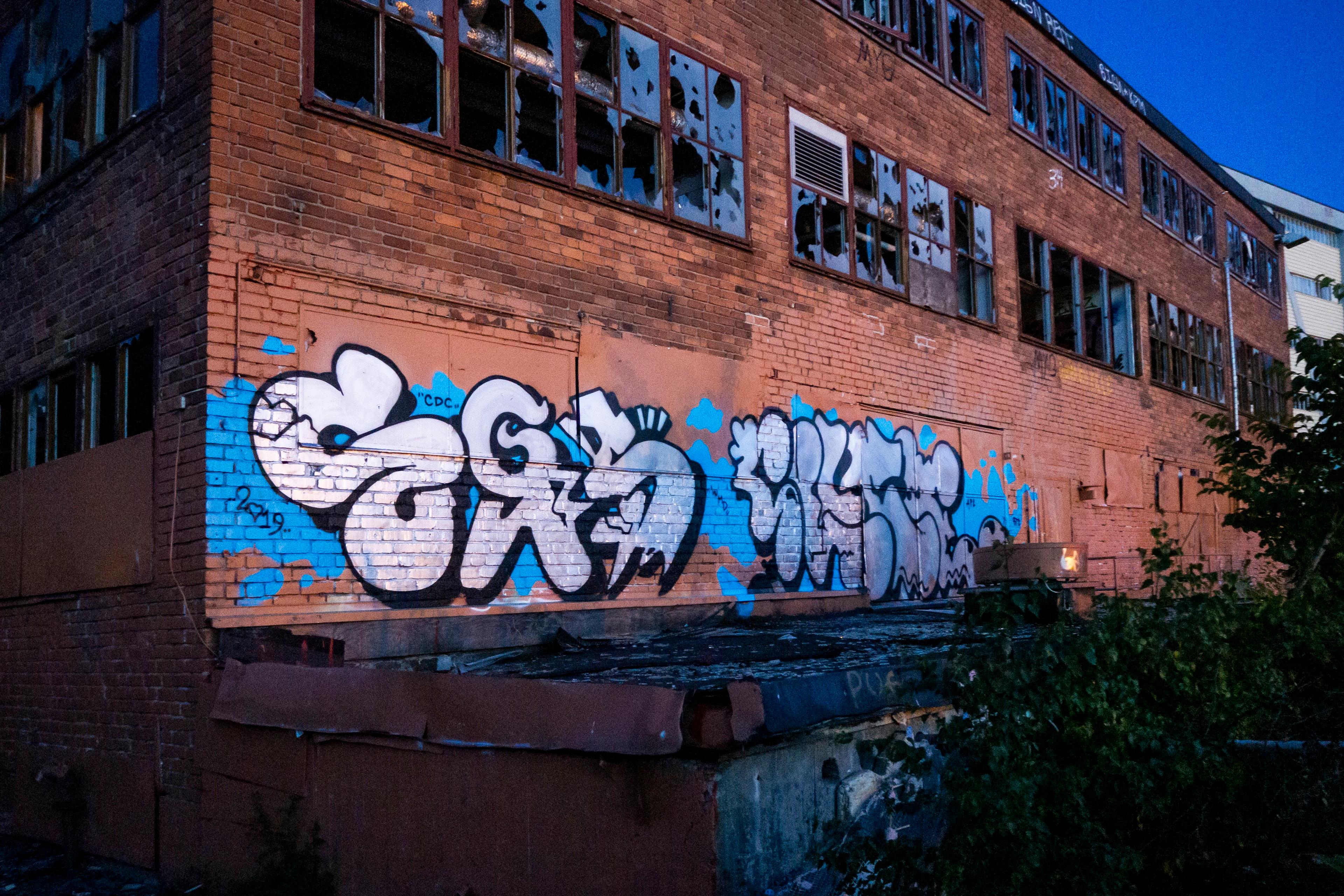 Photo of an abandoned brick building with graffiti on its walls broken windows and overgrown plants in the foreground.