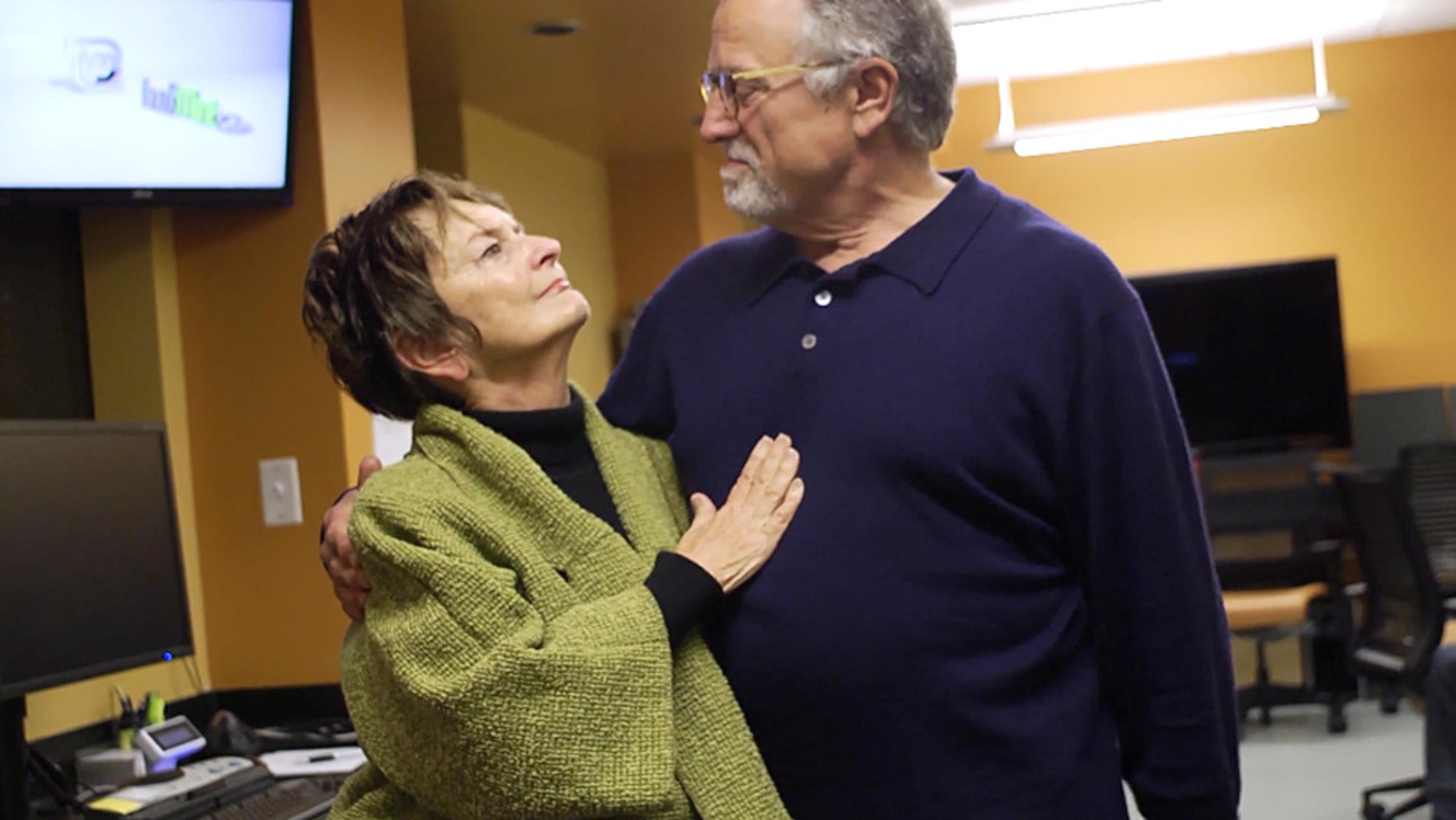 An elderly couple embracing in an office. The woman wears a green shawl, the man wears a navy jumper, they gaze into each other’s eyes.