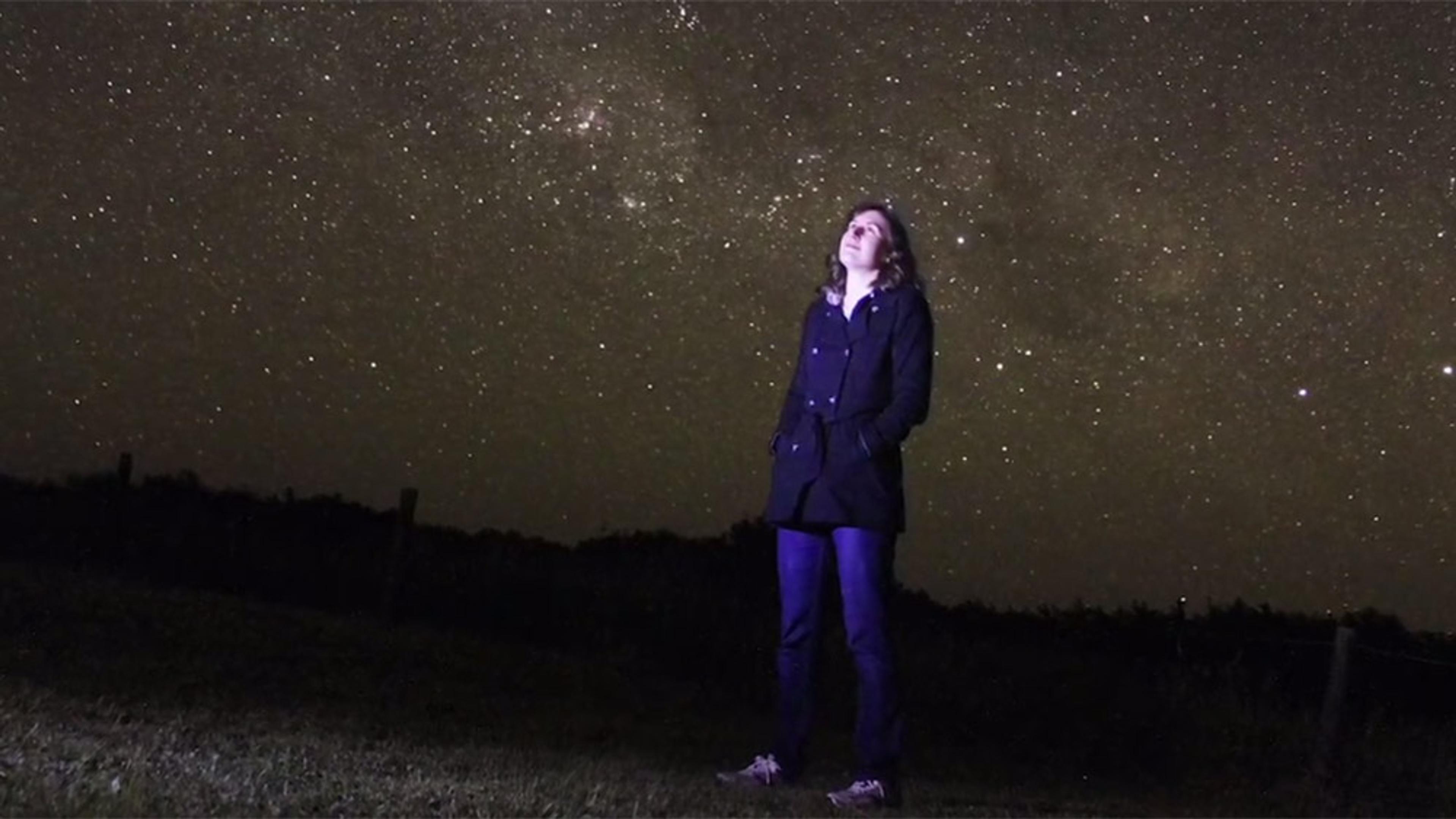 A person standing outdoors at night, looking up at a star-filled sky with hands in pockets, silhouetted against the horizon.