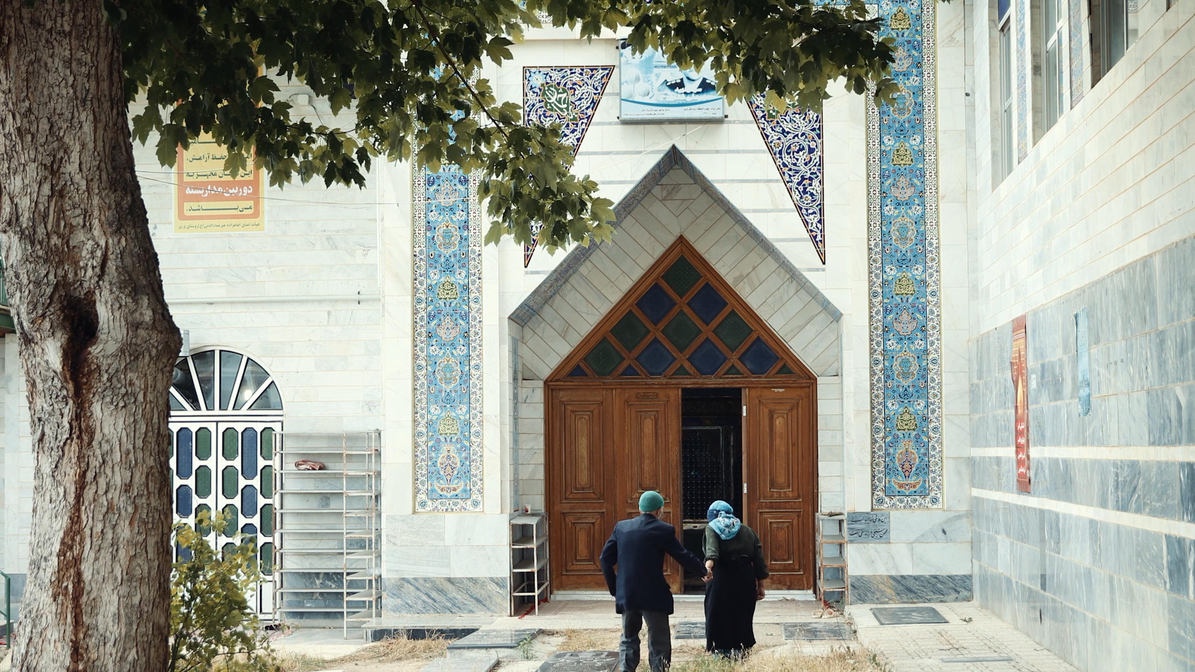 Photo of two people entering a building with ornate blue mosaic tiles and large wooden doors, tree in foreground.
