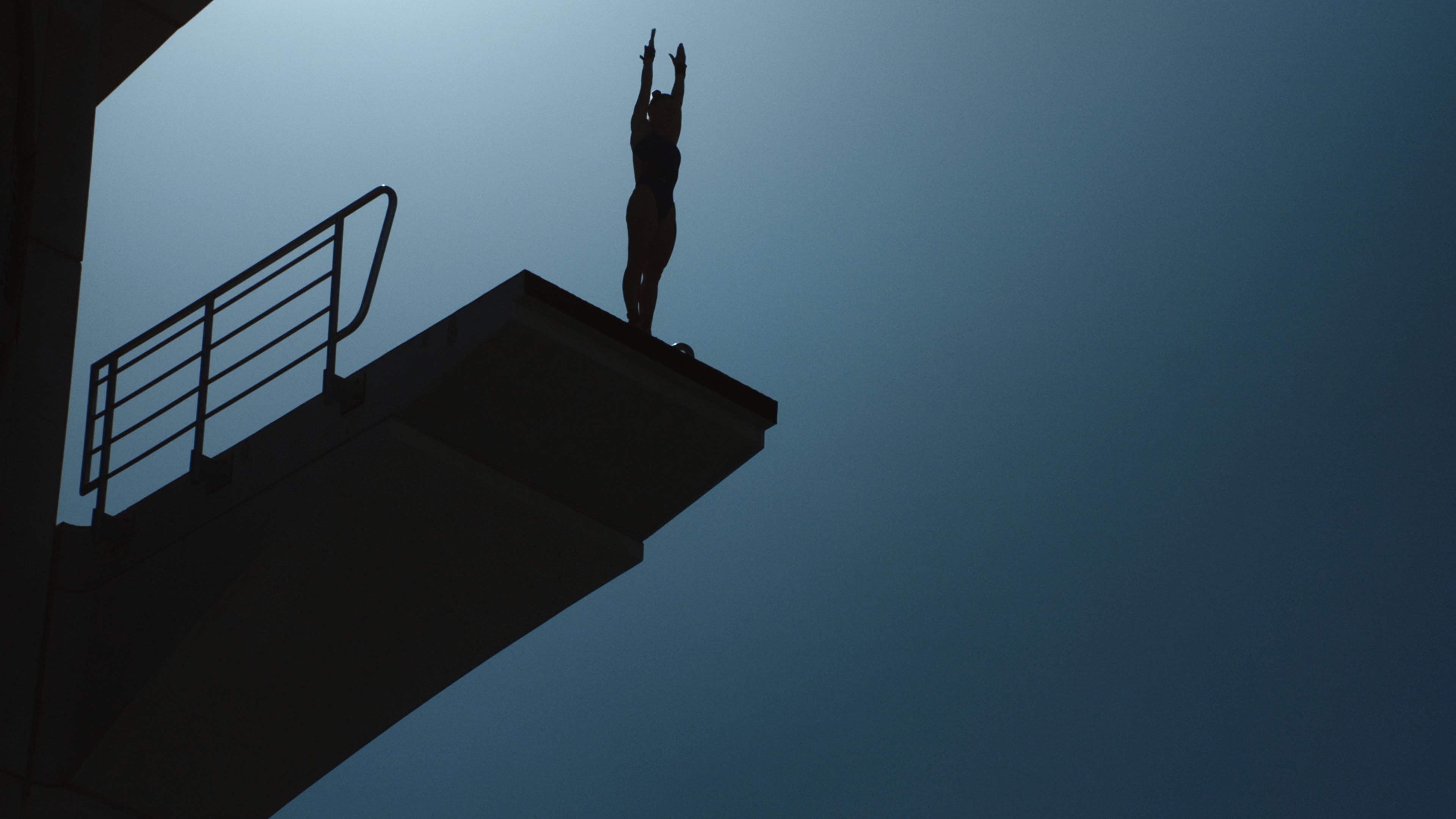 Photo of a silhouetted diver poised on a high diving board against a dark blue sky.
