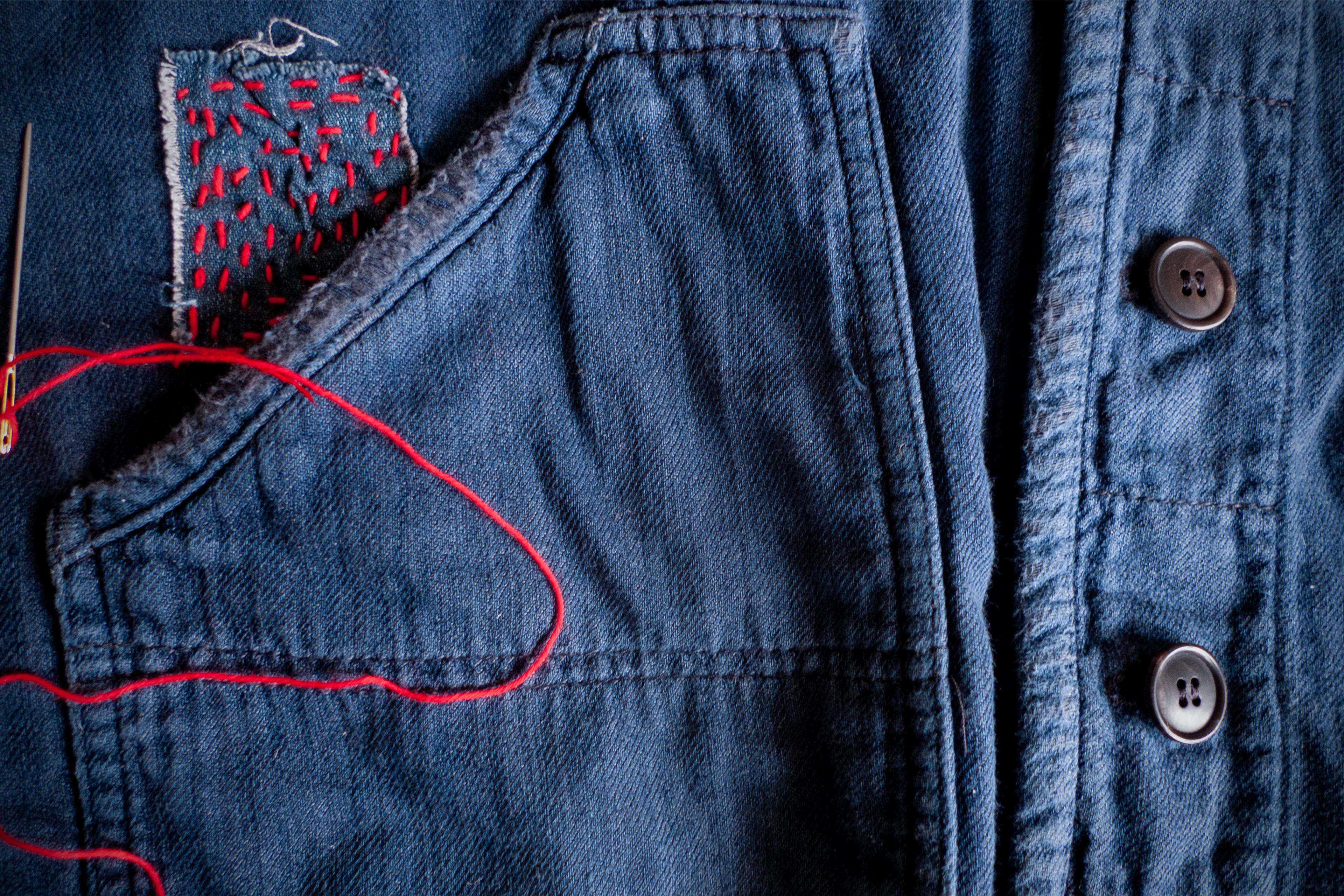 A denim jacket with a patch sewn on with red thread, with a needle with thread resting on the jacket