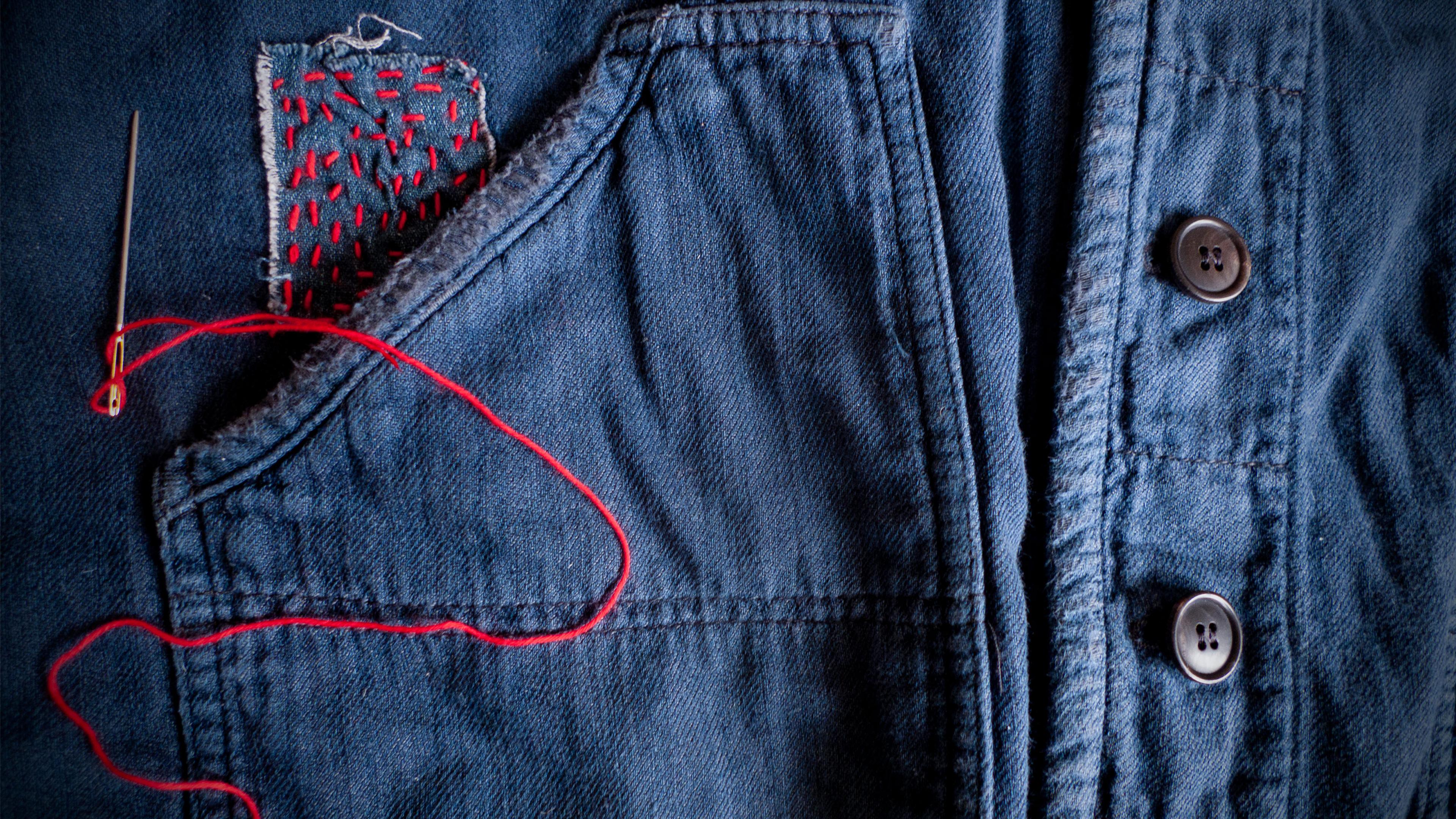A denim jacket with a patch sewn on with red thread, with a needle with thread resting on the jacket