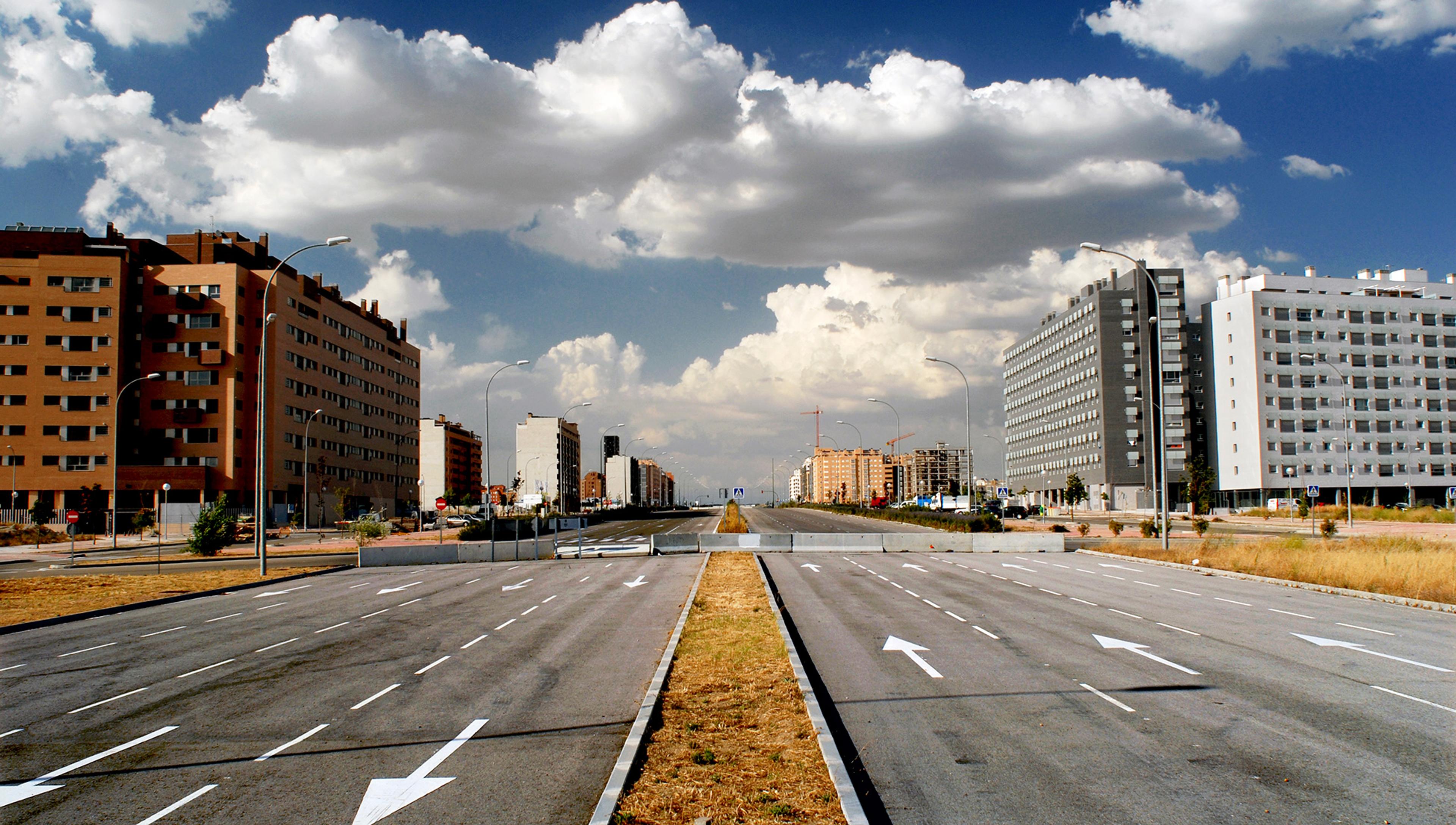 Wide, empty urban road flanked by modern apartment buildings under a partly cloudy sky.