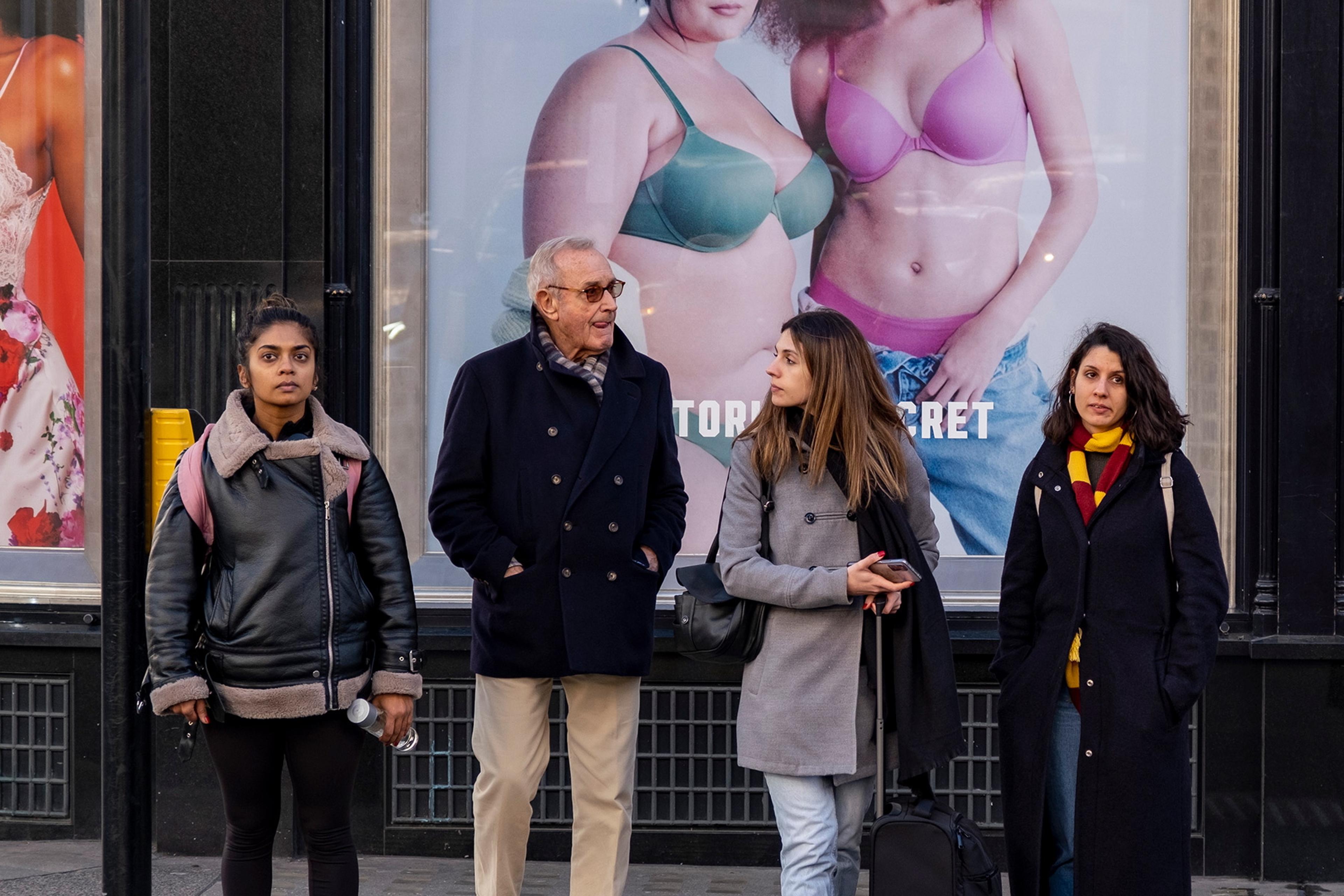 Photo of four people waiting at a crossing in front of a shop with large fashion advertisements in the background.