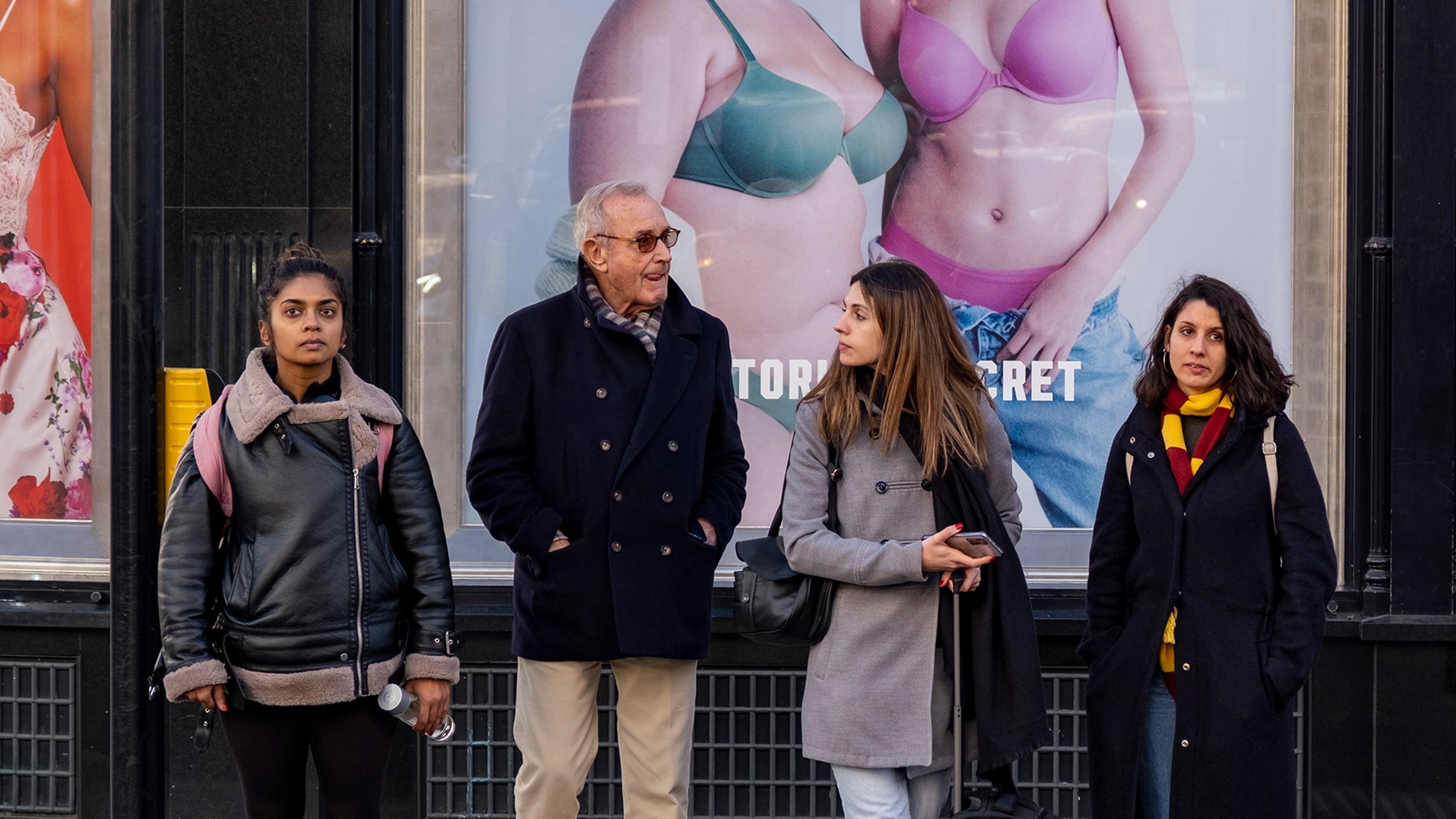 Photo of four people waiting at a crossing in front of a shop with large fashion advertisements in the background.