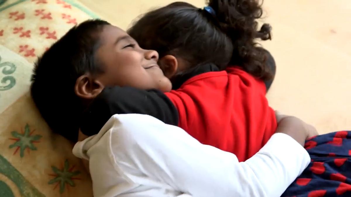 Two children hugging on the floor, one in a white shirt smiling with eyes closed while the other wears a red jacket.