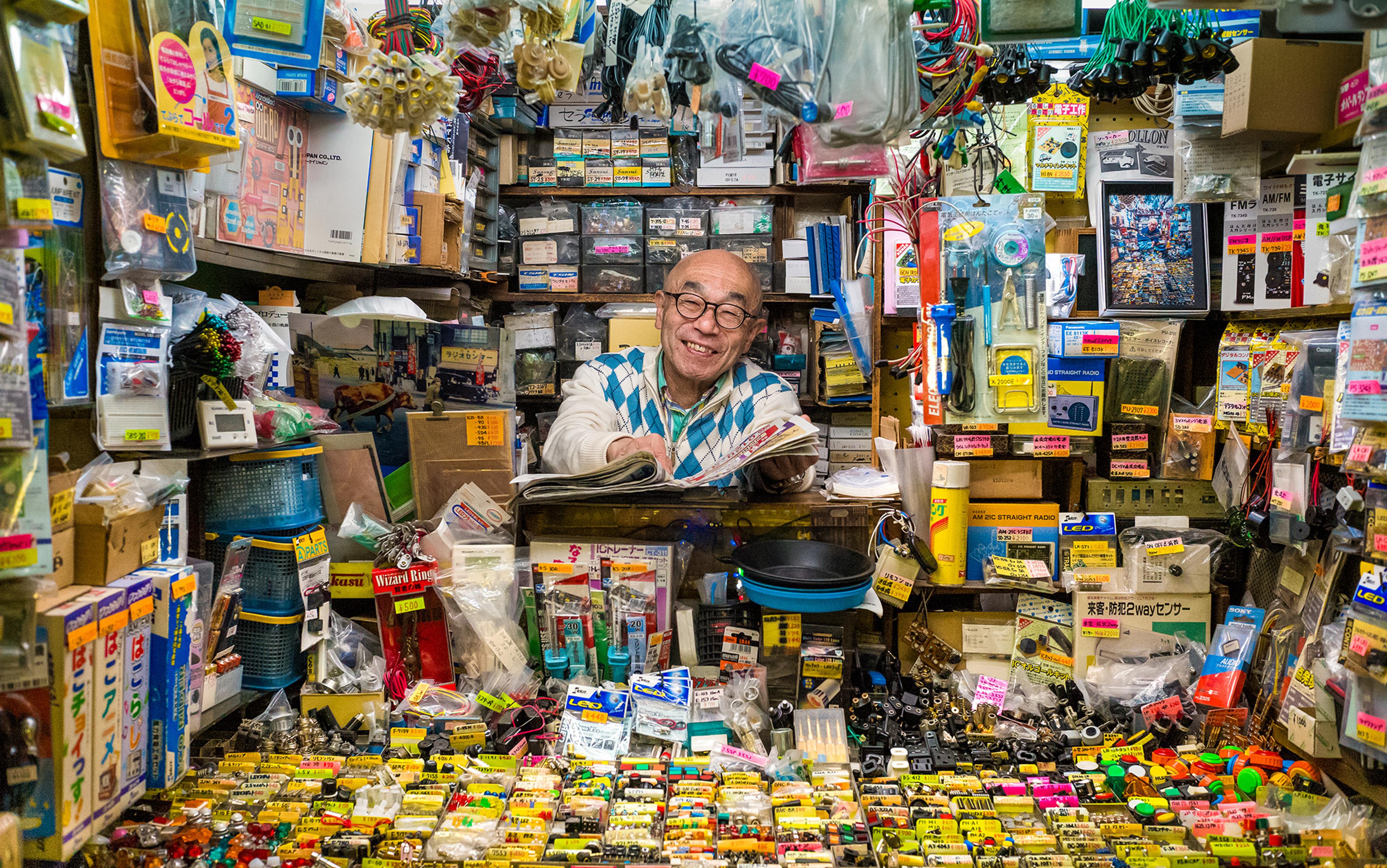 A smiling elderly man in a cluttered electronics shop with various gadgets and boxes surrounding him.