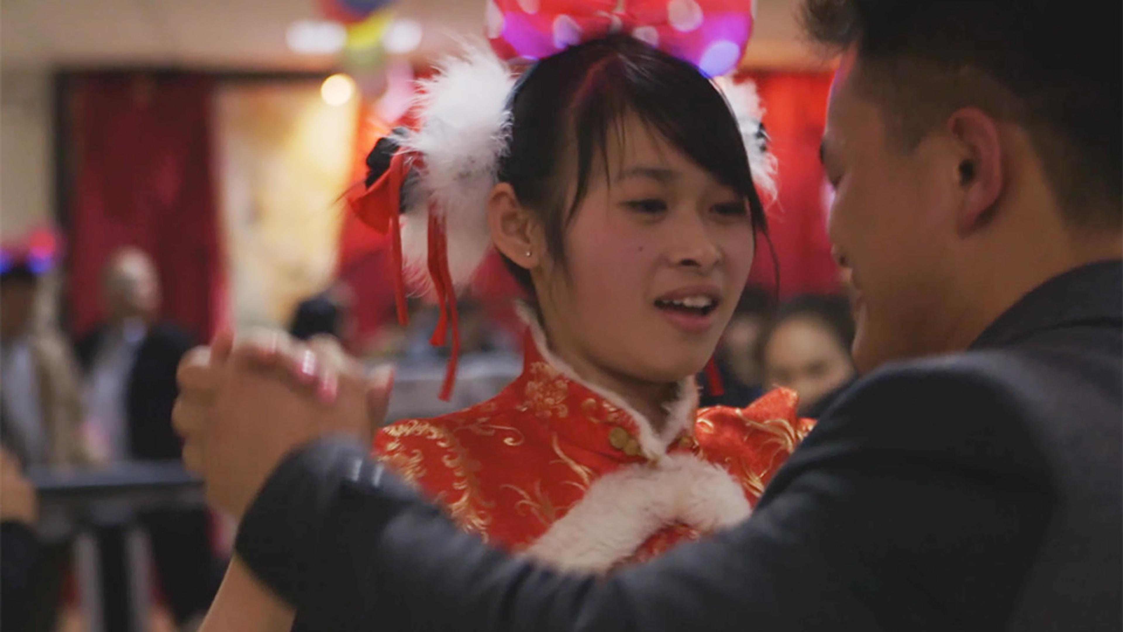A woman in a red Chinese outfit dancing with a man; she wears festive headgear with white feathers.