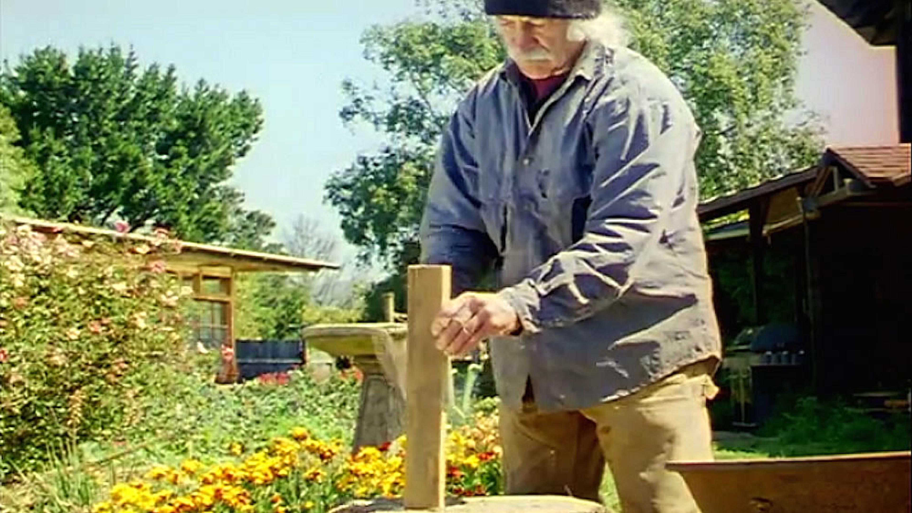 An elderly man working with wood outside. He is wearing a beanie and jacket. Garden and building are in the background.