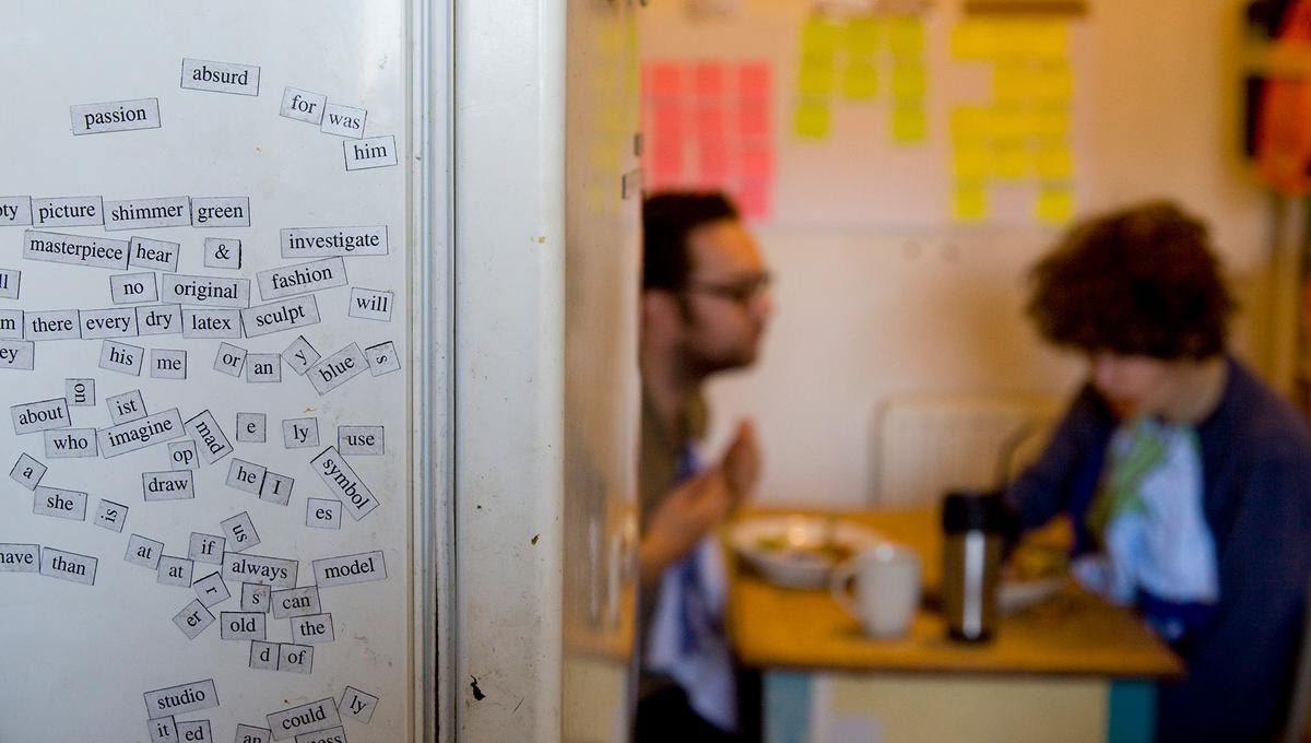 A fridge covered in word magnets in the foreground next to two people having a meal at a table with sticky notes on the wall blurred in the background.