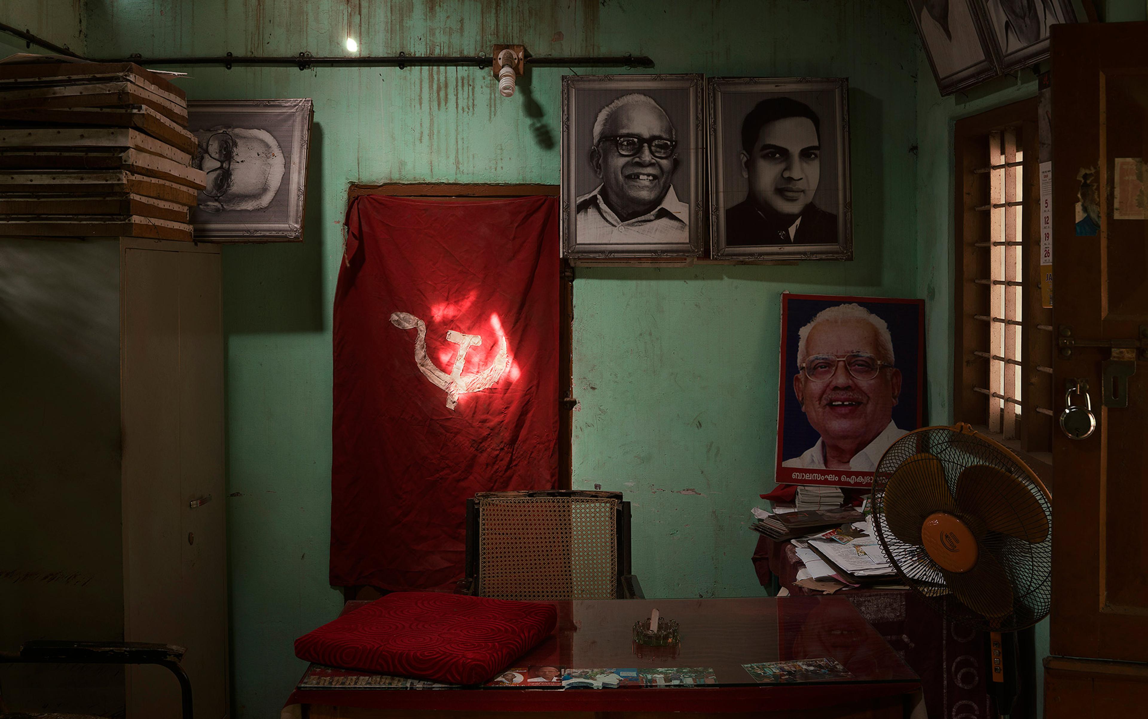 An office with framed portraits of three men on a green wall, a red flag with a hammer and sickle, a desk, and a table fan.