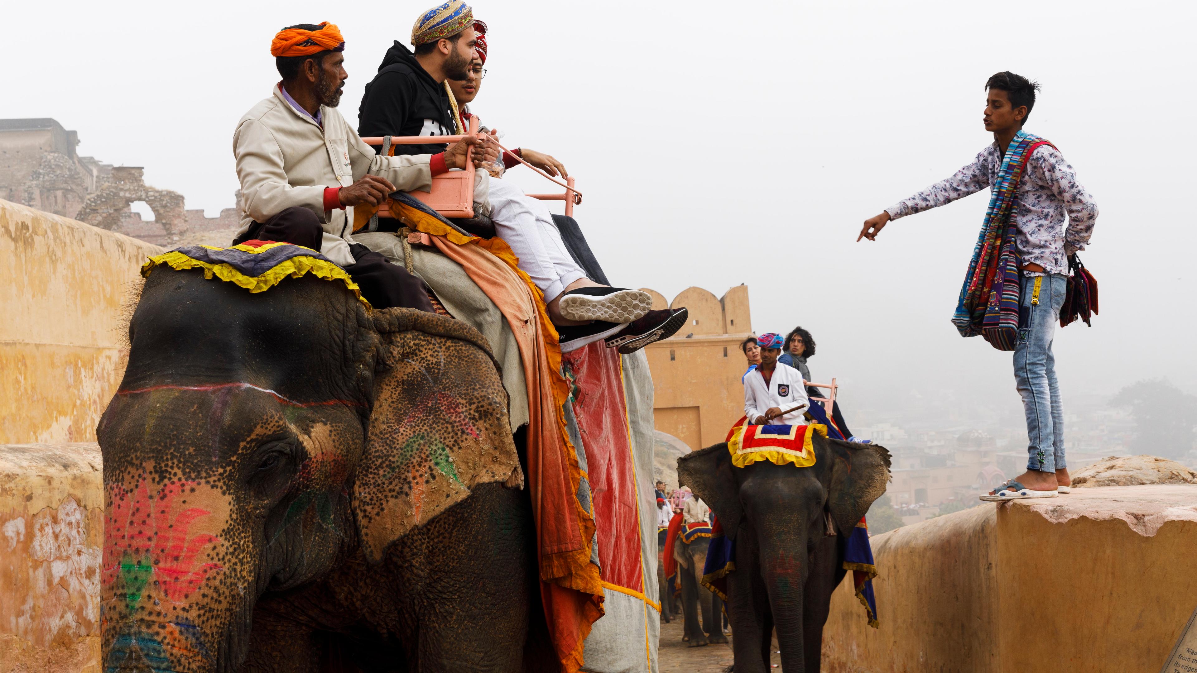 Photo of people riding brightly adorned elephants on a stone path with a vendor standing nearby pointing.