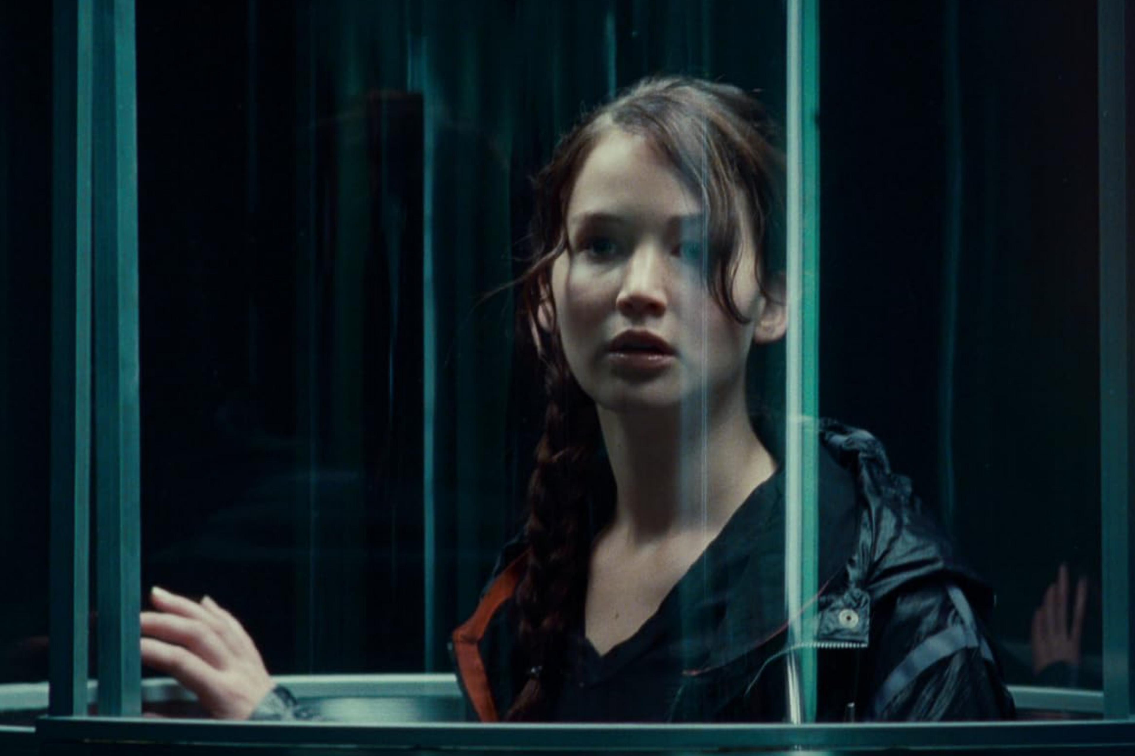 A film still of a woman, Katniss Everdeen from The Hunger Games movie franchise, with a braid standing in a transparent elevator looking out with a thoughtful expression.
