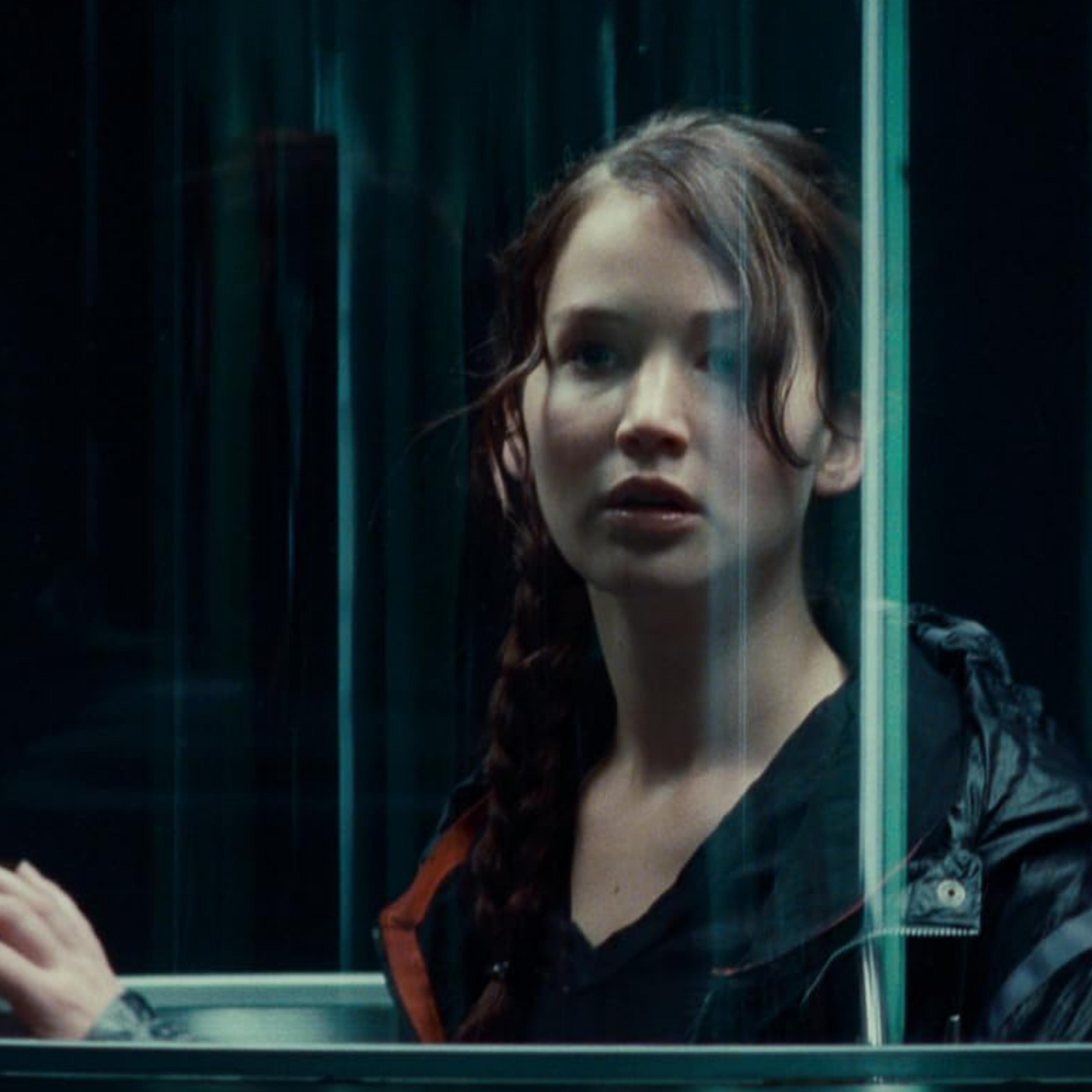 헝거게임 영화 시리즈의 캐트니스 에버딘이라는 여성이 머리를 땋고 투명한 엘리베이터 안에 서서 생각에 잠긴 표정으로 바깥을 바라보는 장면입니다.<BR>