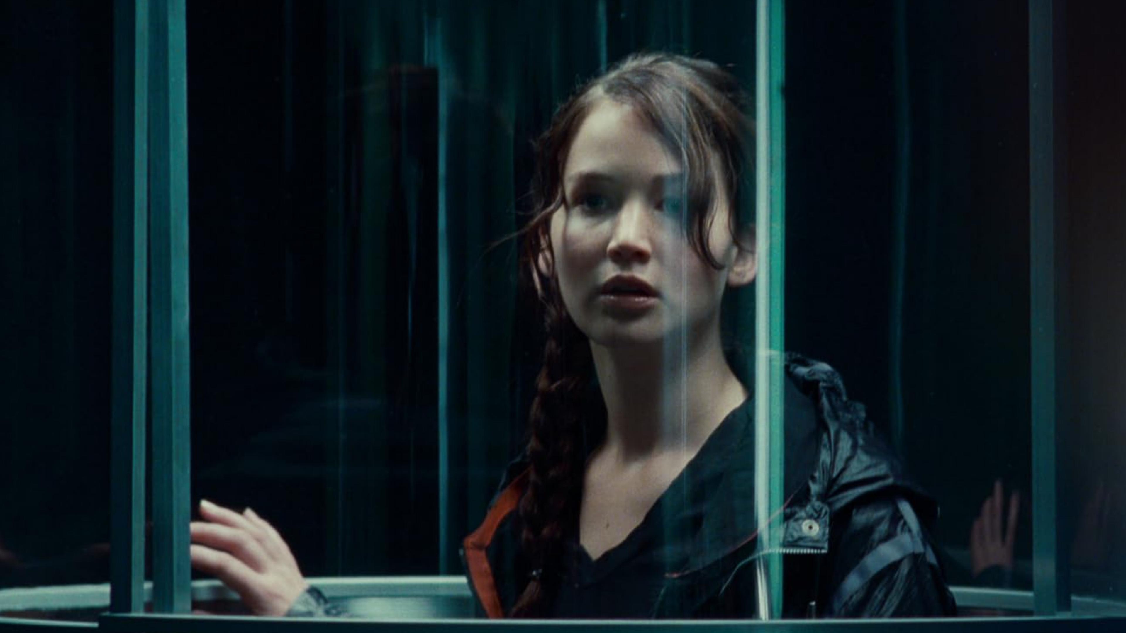 A film still of a woman, Katniss Everdeen from The Hunger Games movie franchise, with a braid standing in a transparent elevator looking out with a thoughtful expression.