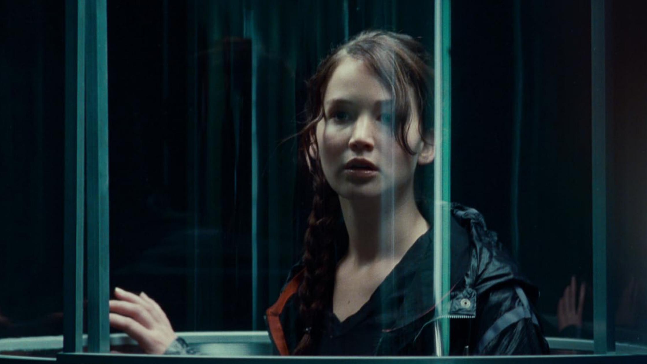 A film still of a woman, Katniss Everdeen from The Hunger Games movie franchise, with a braid standing in a transparent elevator looking out with a thoughtful expression.