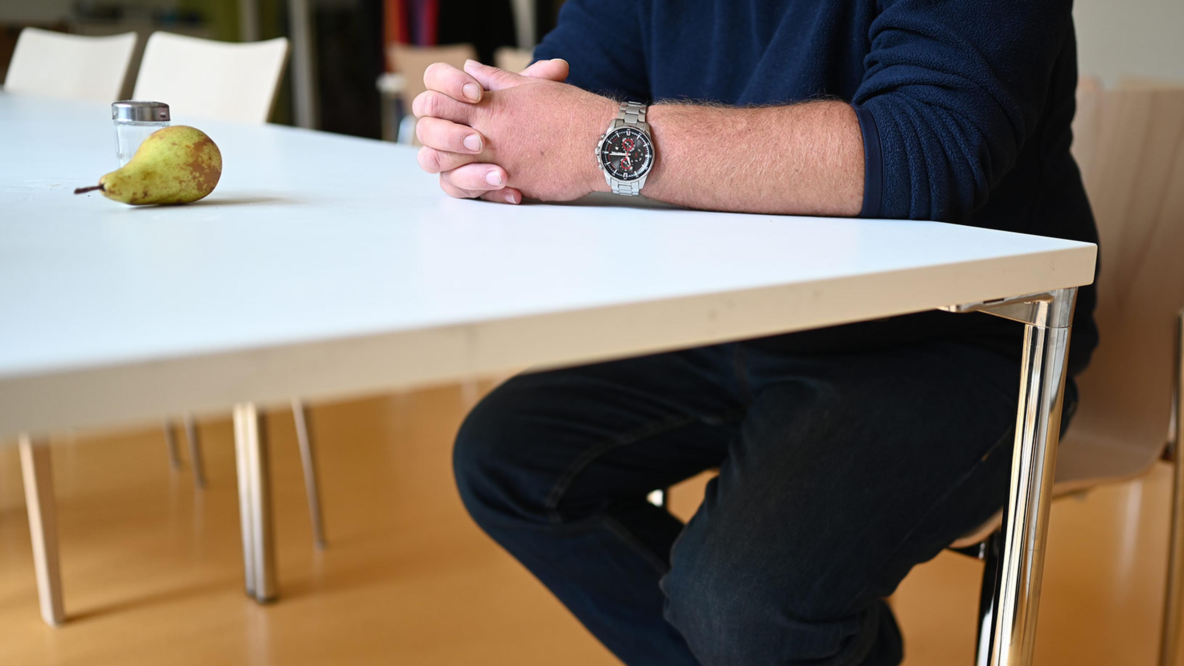 A man seated at a white table with hands folded, wearing a watch. A pear and a salt shaker on the table, chairs in background.
