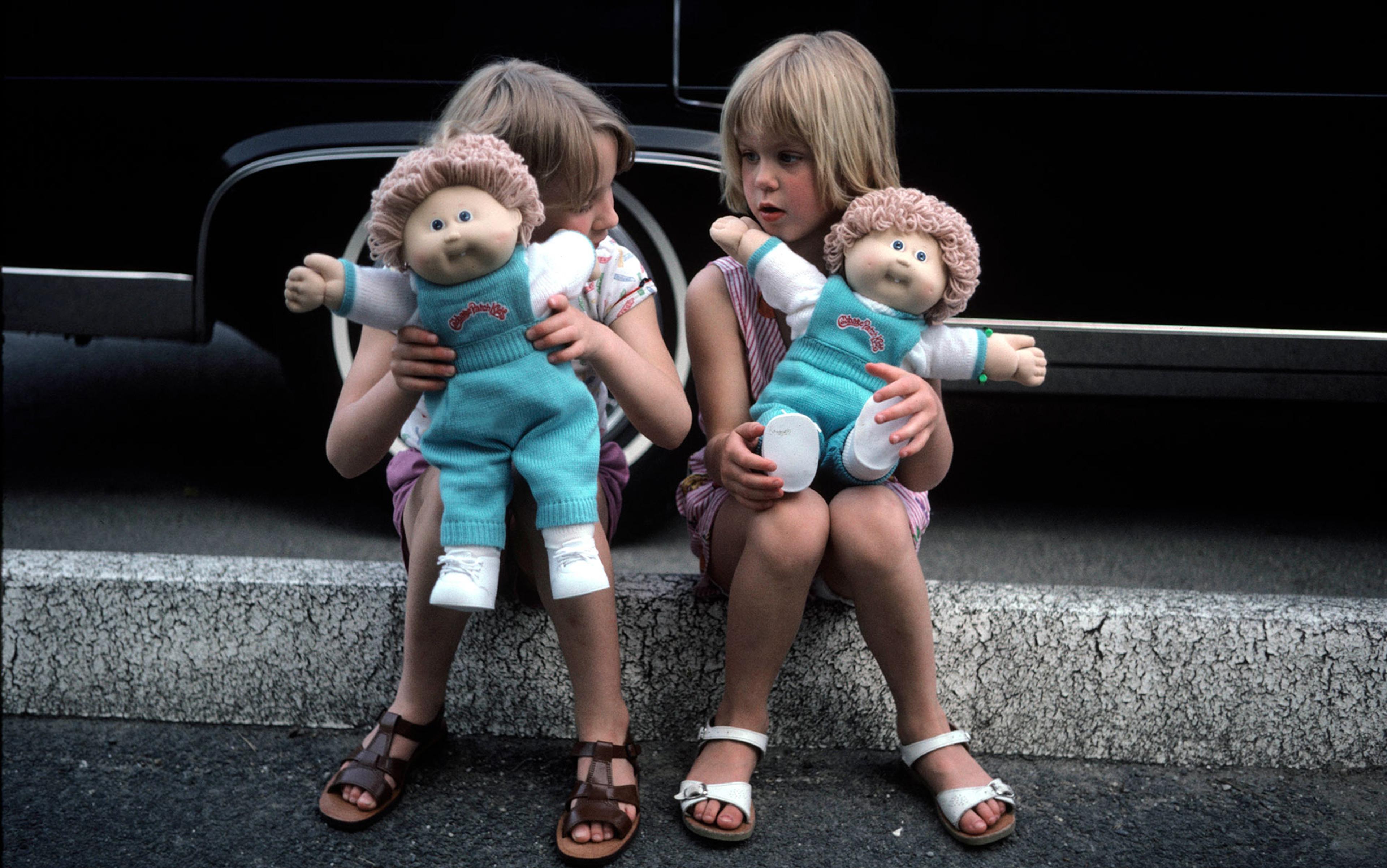 Two children sitting on a curb, each holding a Cabbage Patch Kid doll, with a black car in the background.
