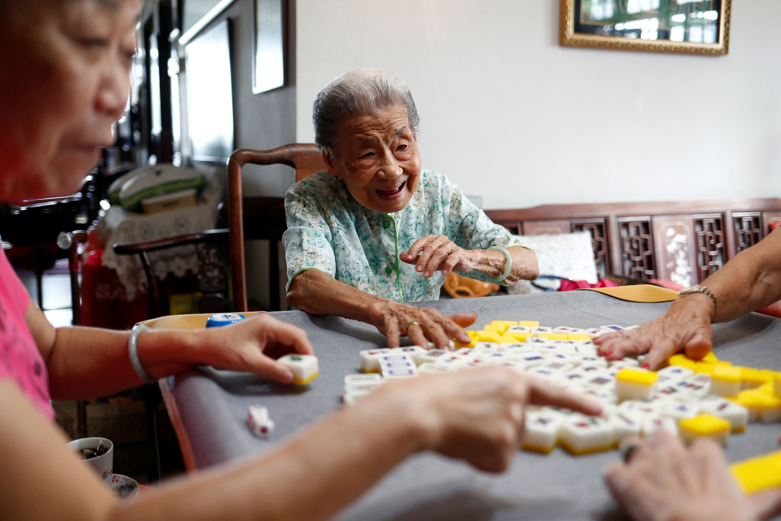 Photo of elderly women playing Mahjong at a table, laughing and engaged in the game in a cosy indoor setting.
