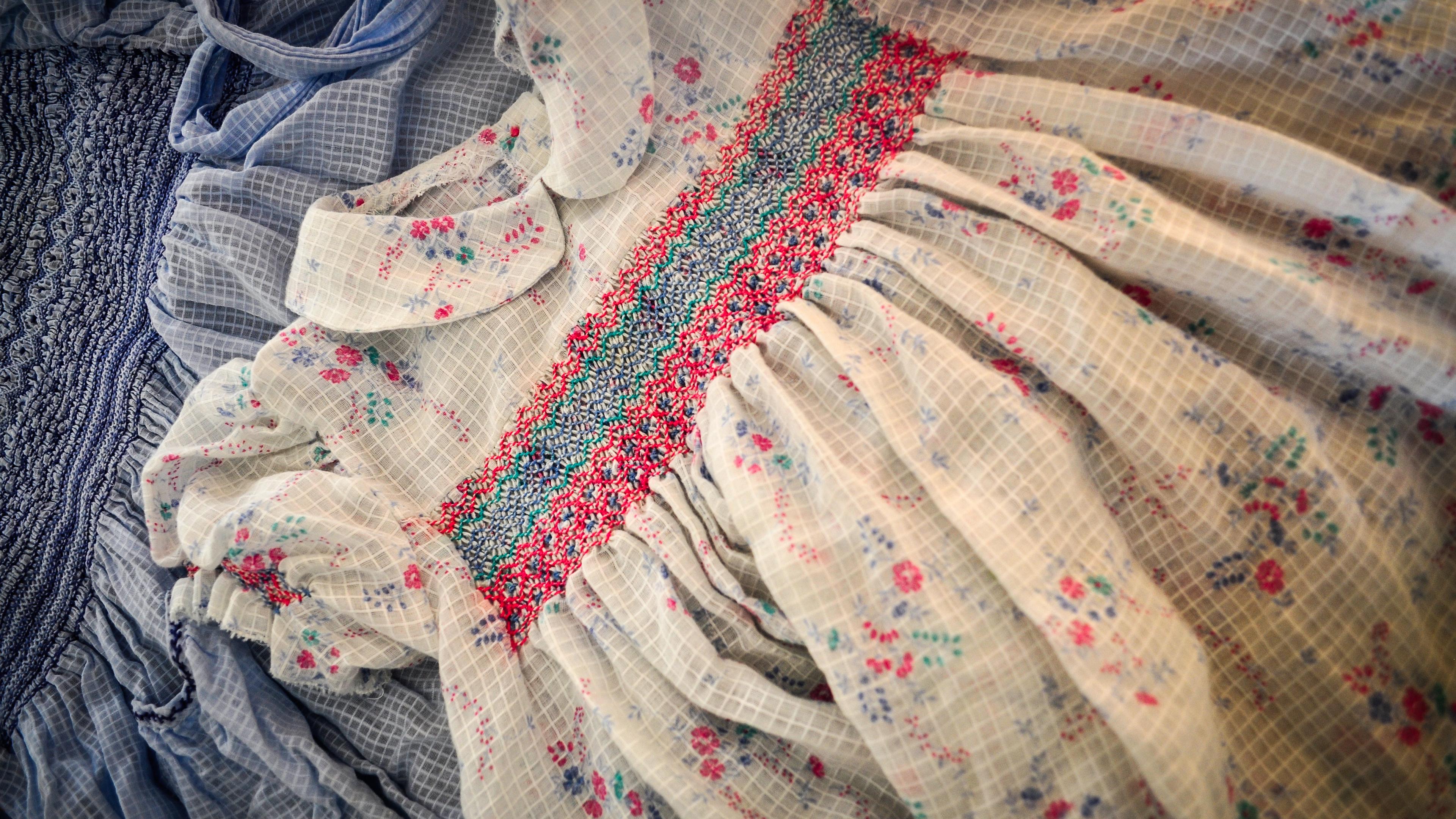Two children’s dresses lying flat, one blue the other white with floral patterns and colourful smocking.