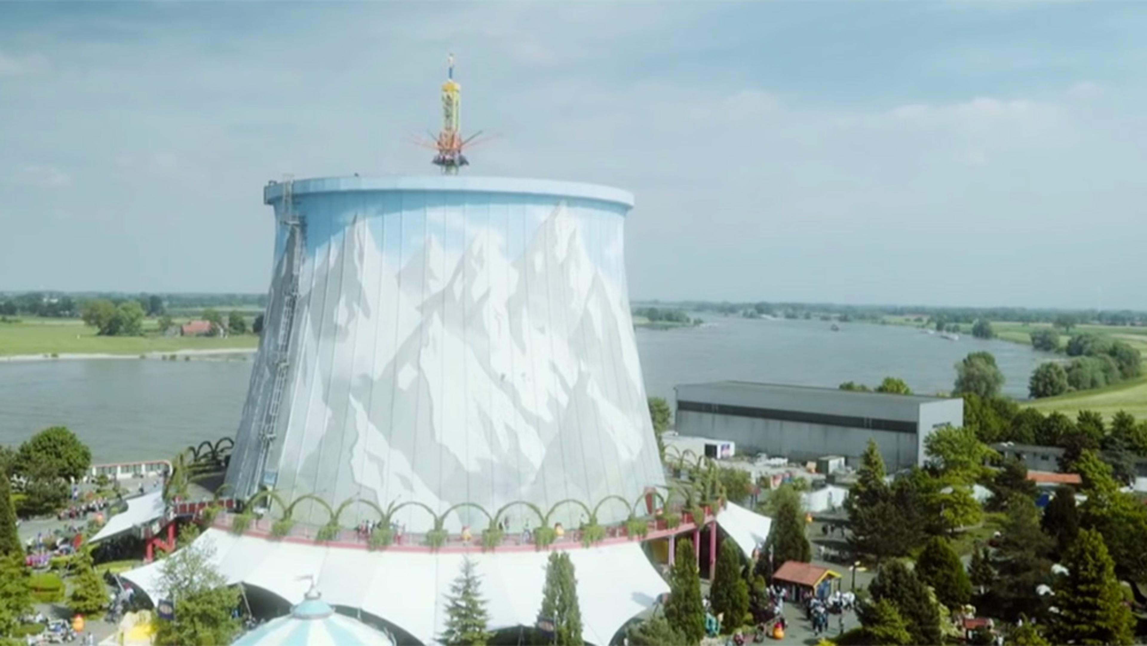 A water ride built around a large tower with a mural of snowy mountains, by a river with surrounding greenery.