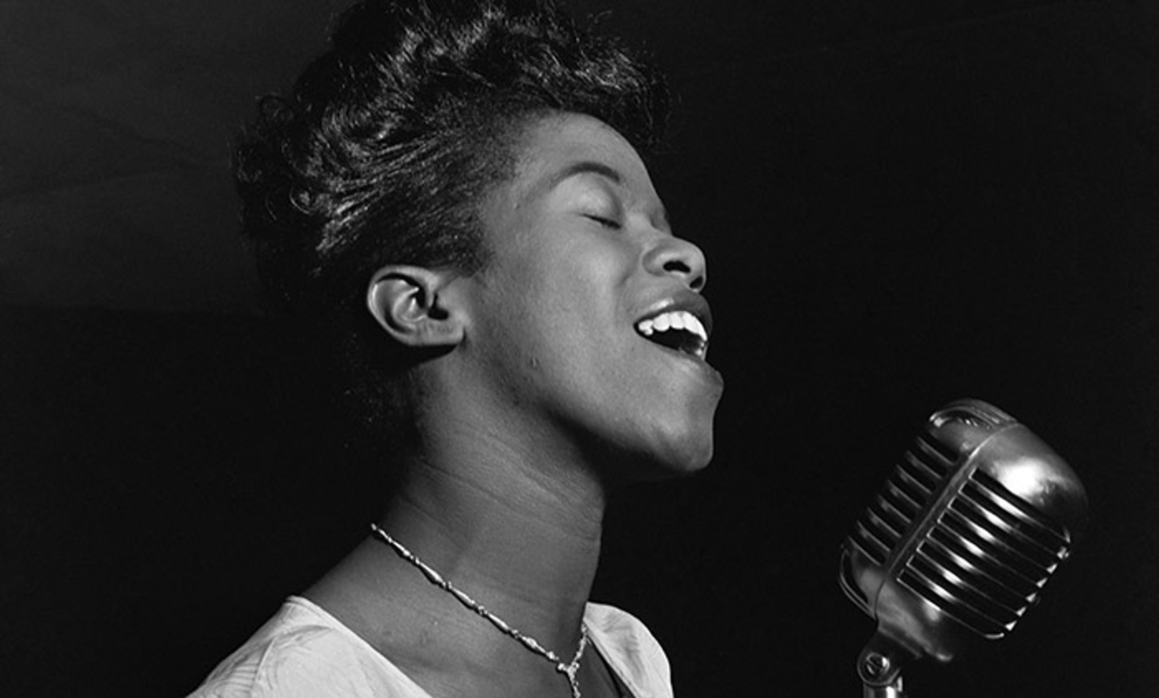 Black and white photo of a Black woman singing passionately into a vintage microphone while wearing a necklace.