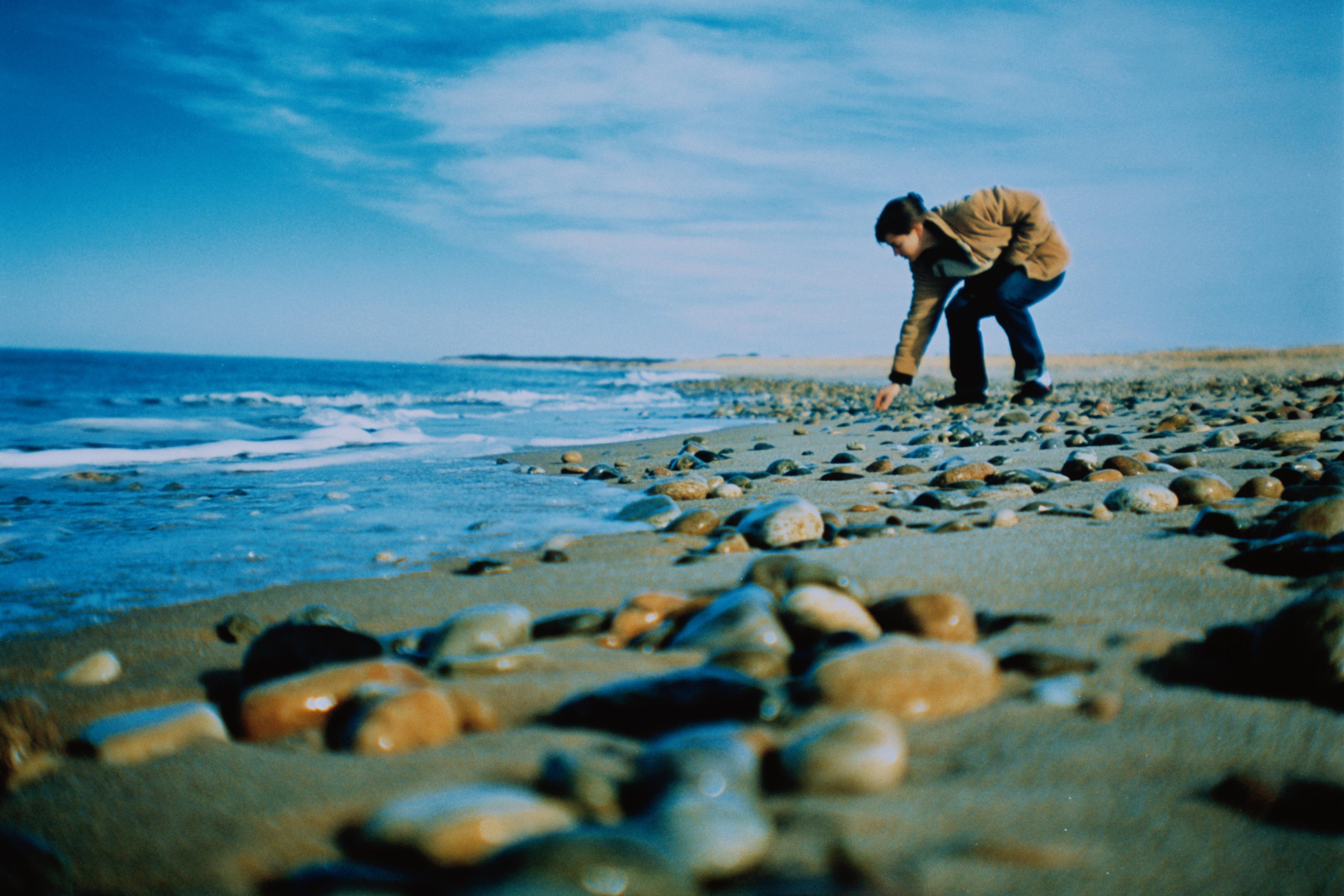 Photo of a person picking up stones on a pebbly beach with a blue sky and ocean waves in the background.