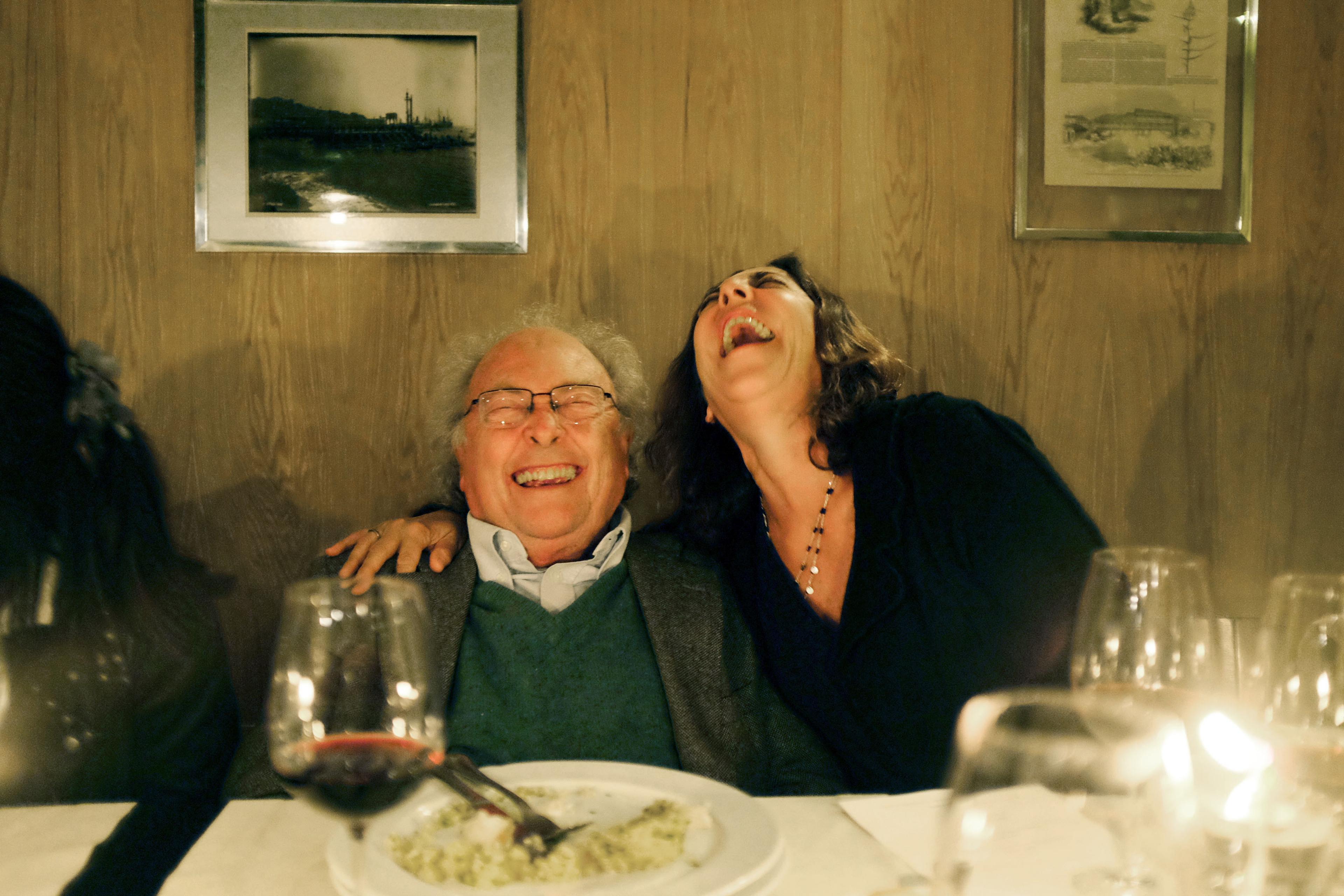 Photo of two people at a dinner table laughing heartily with wine glasses and a plate in the foreground.